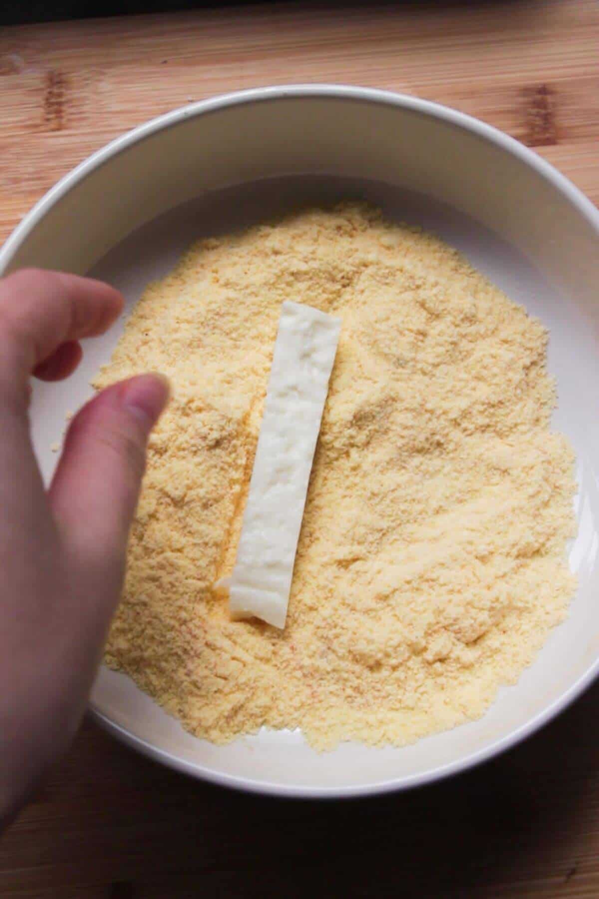 Hand coating a piece of halloumi in polenta in a small white bowl.