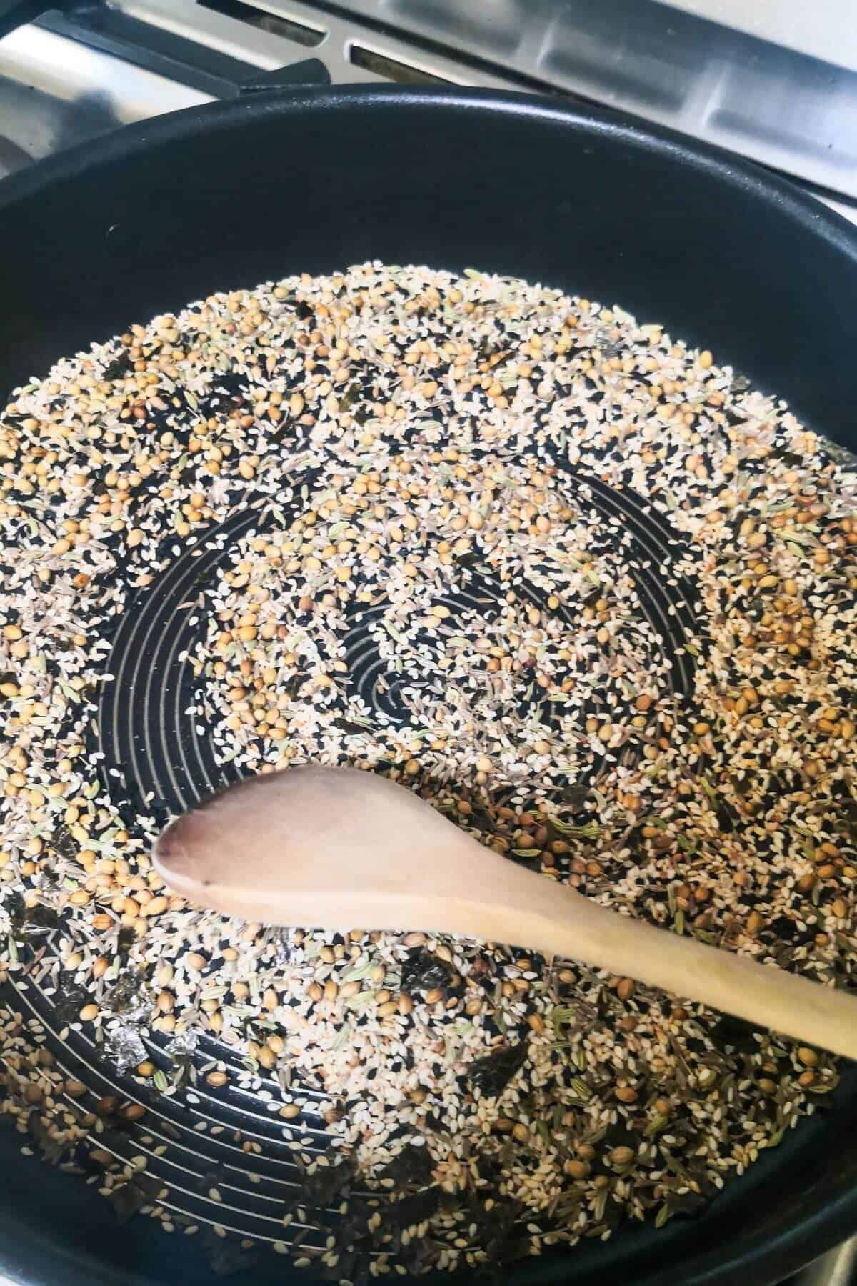 Seeds being toasted in a large frying pan with a wooden spoon stirring them.
