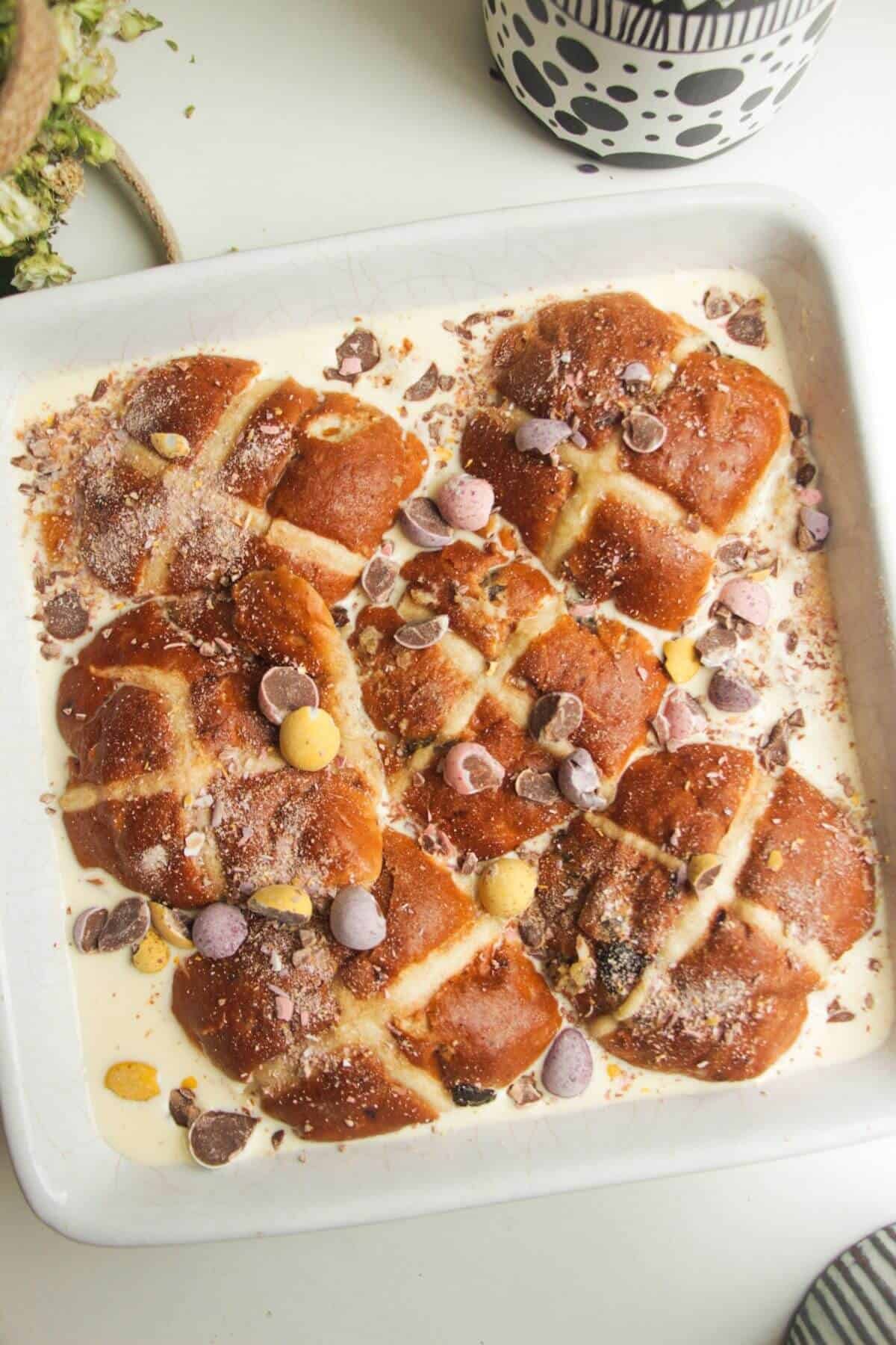 Hot cross buns layered with custard and mini Easter eggs in a square baking dish.