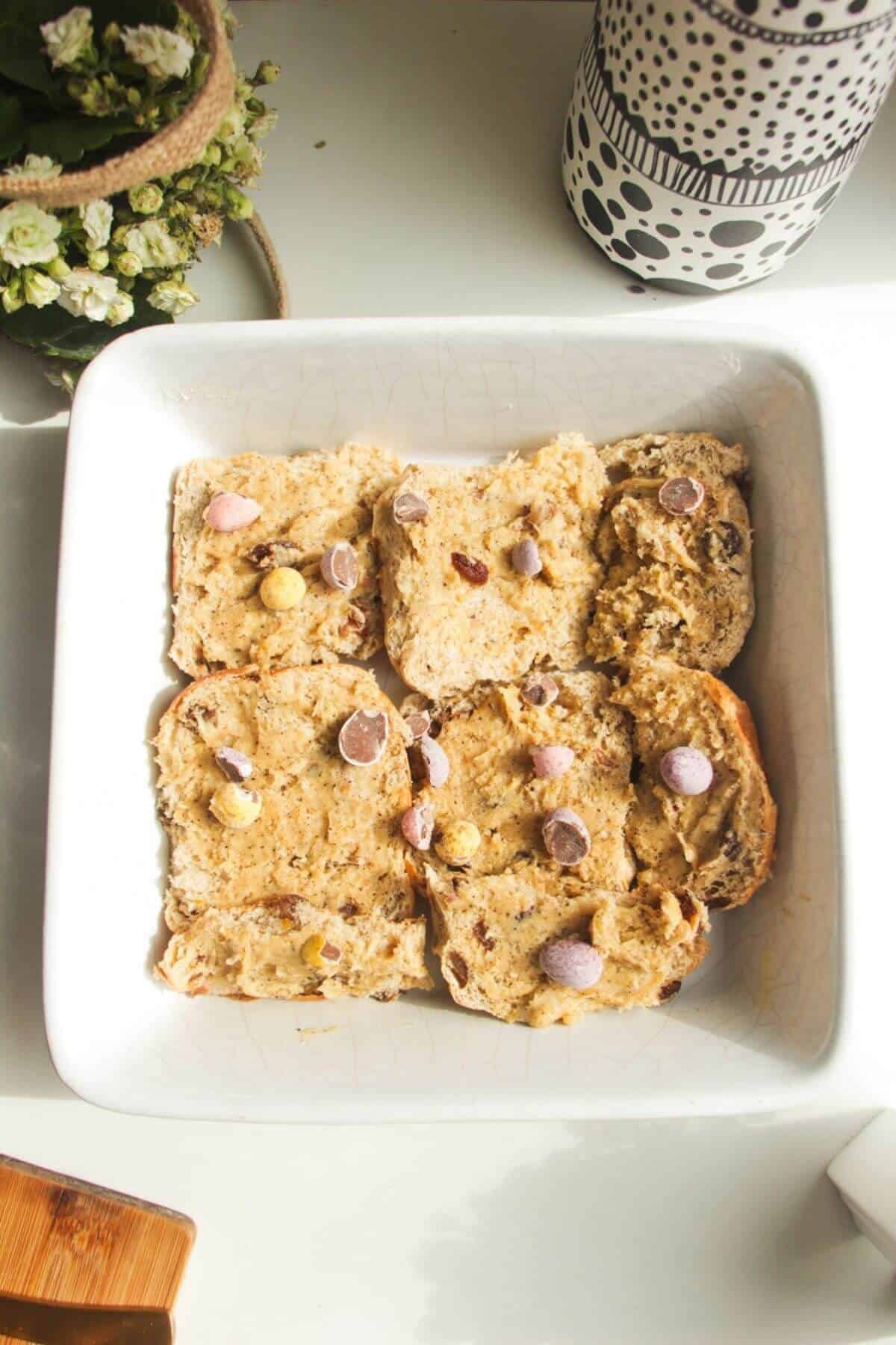 Buttered hot cross buns scattered with min Easter eggs in a baking dish.