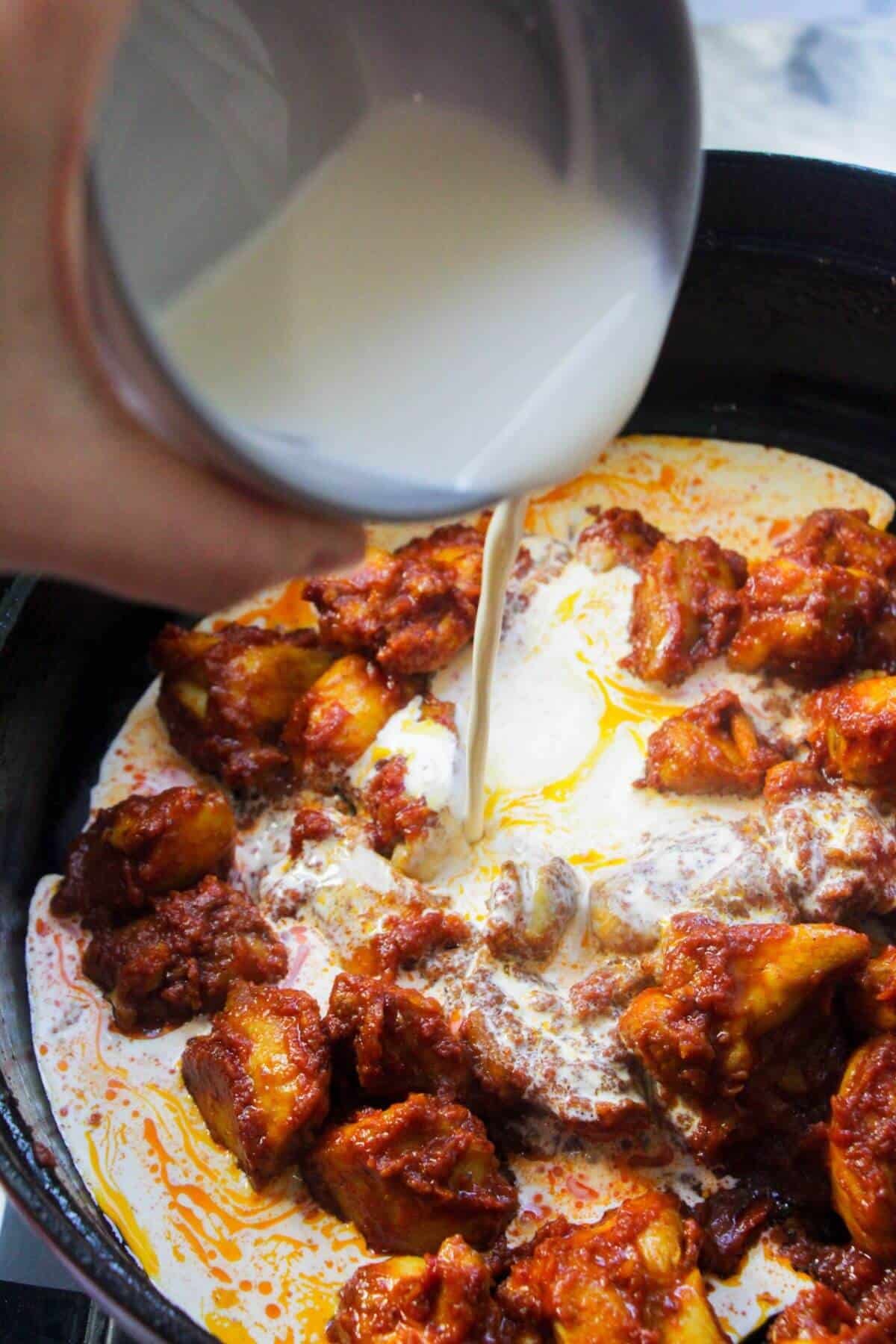 Cream being poured into a large black pan with tomato coated chicken thigh pieces.