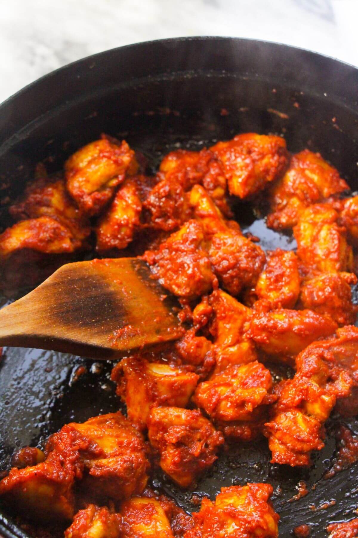 Tomato coated chopped chicken thighs being cooked in a large black pan with a wooden spatula stirring.