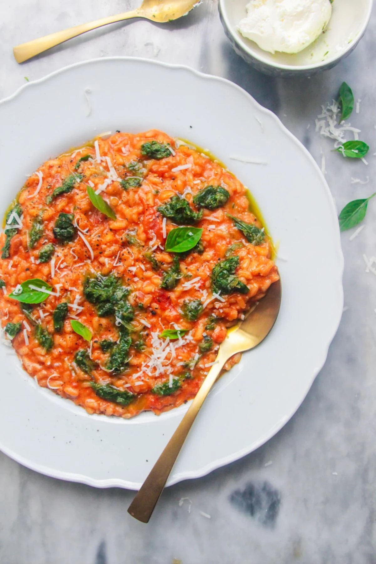 Tomato mascarpone risotto in a large white plate with dollop of green pesto on top, with a gold spoon in the risotto and bowl of mascarpone in the background.