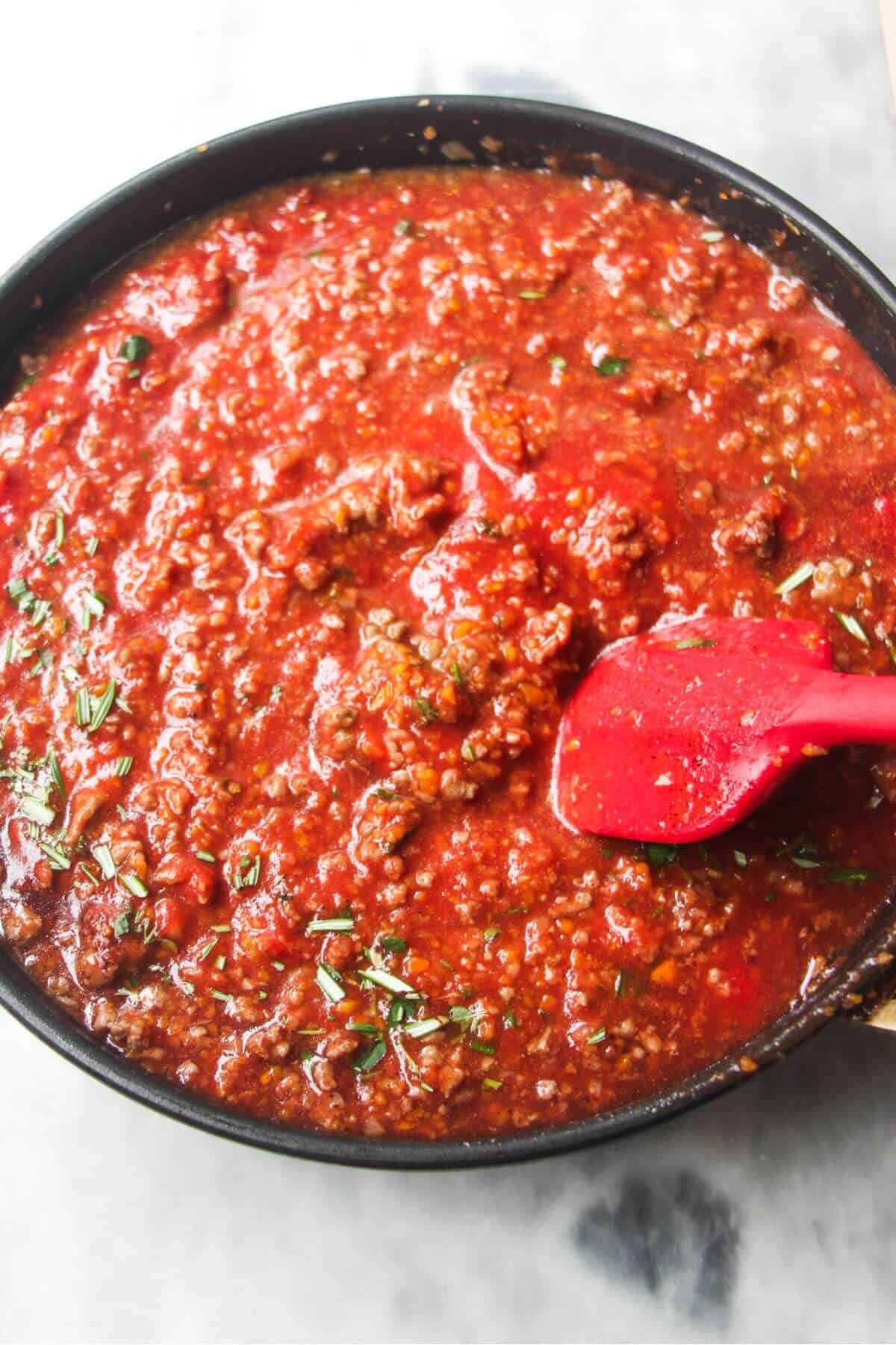 Red spatula stirring tomatoes, red wine and herbs through ground beef in a small black pan.