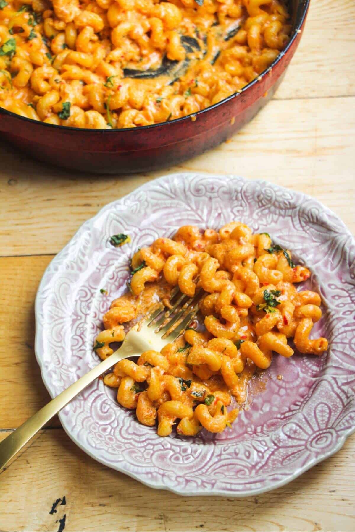 Spicy mac and cheese on a small pink plate with a skillet with more mac and cheese in the background.