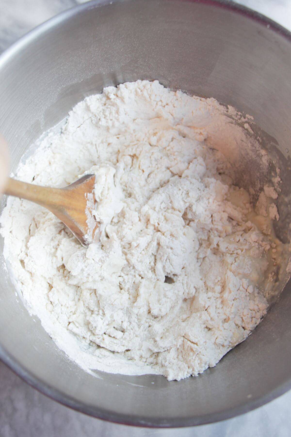 A wooden spatula mixing flour, milk, yeast and salt in a large silver mixing bowl.