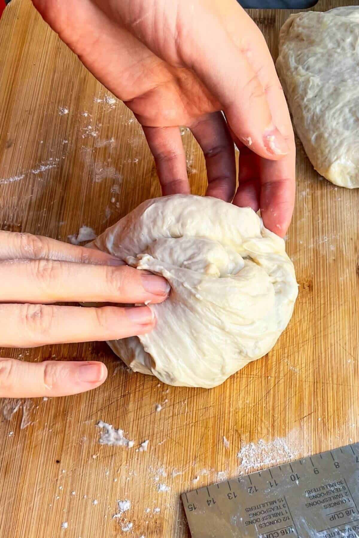 Hands folding pizza dough to form a ball on a wooden board.