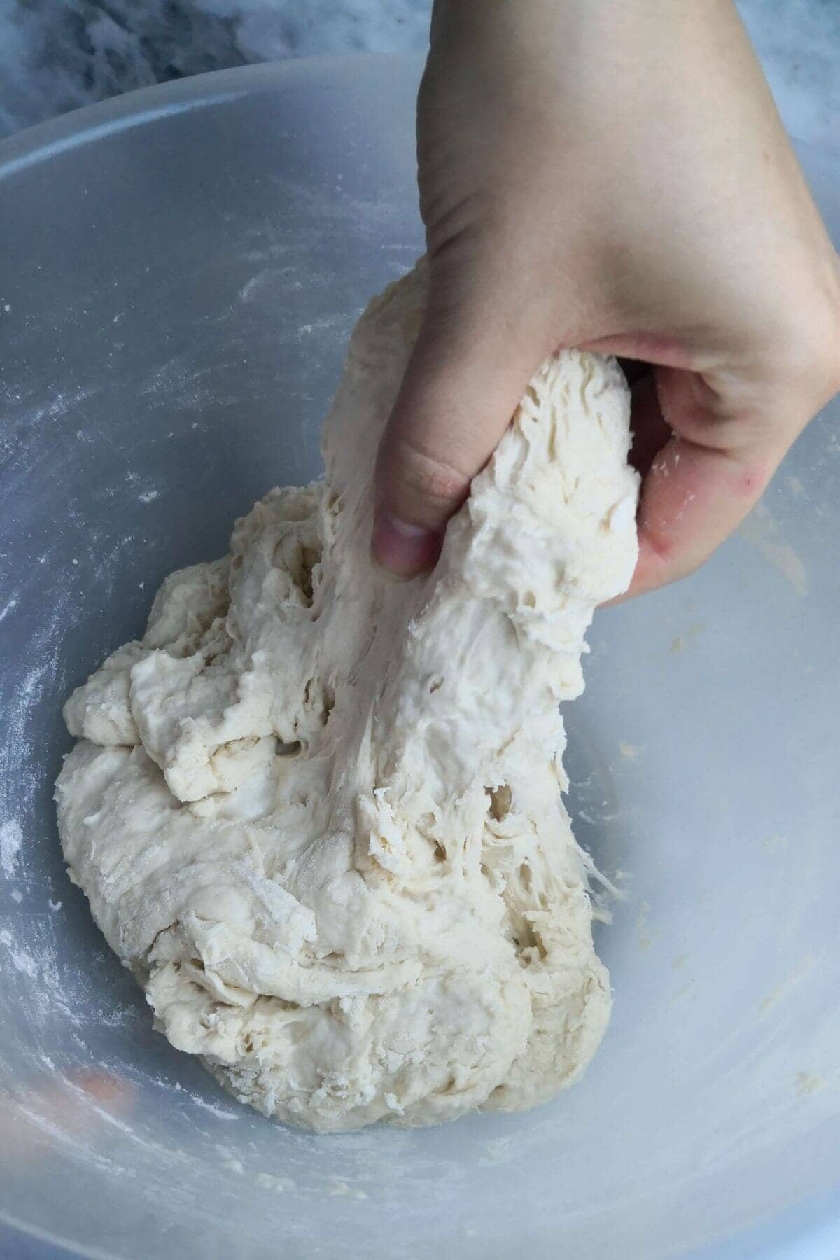 A hand pulling up pizza dough in a large, clear mixing bowl.