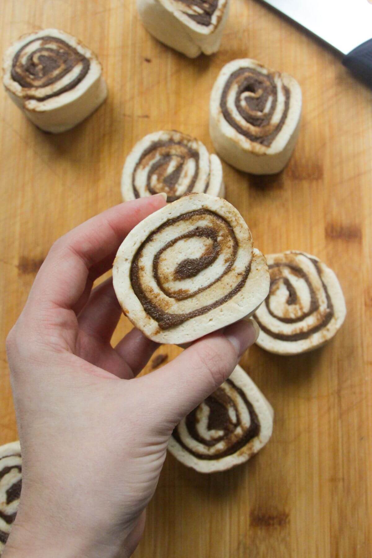 Hand holding up a cinnamon bun after being sliced with more unbaked rolls in the background.