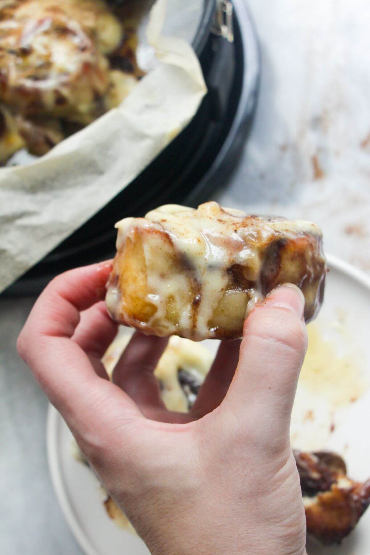 Hand holding up a pumpkin spice cinnamon roll with plate of more rolls in the background.
