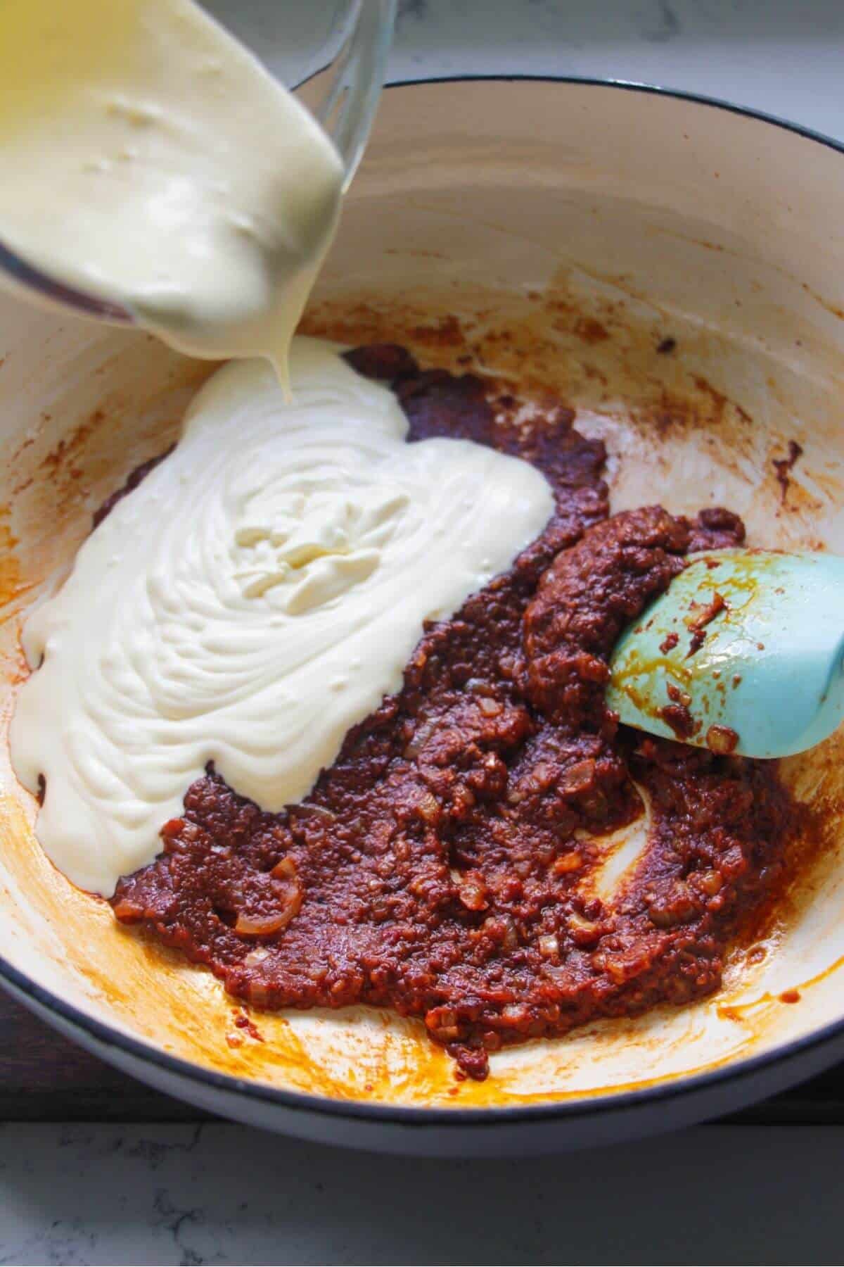 Pouring cream into harissa tomato sauce in a large white skillet.