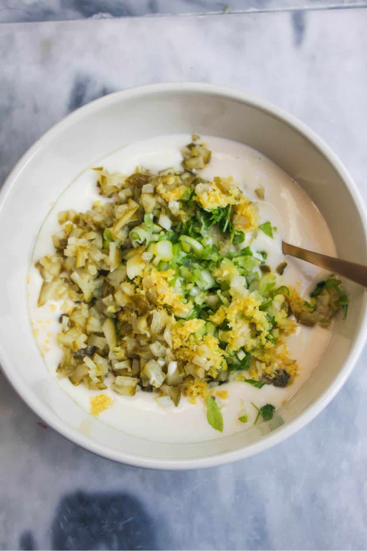 Chopped capers, gherkins, lemon zest on top of aioli in a small white bowl.