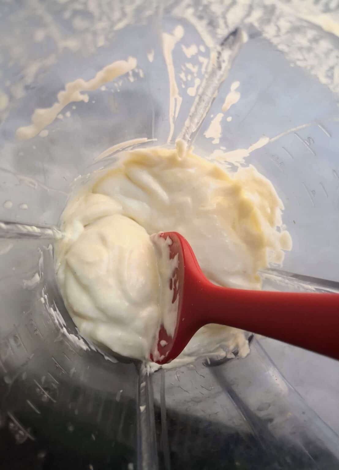White vegan garlic aioli in a clear sided blender with a red spoon scooping some up, looking down from above.