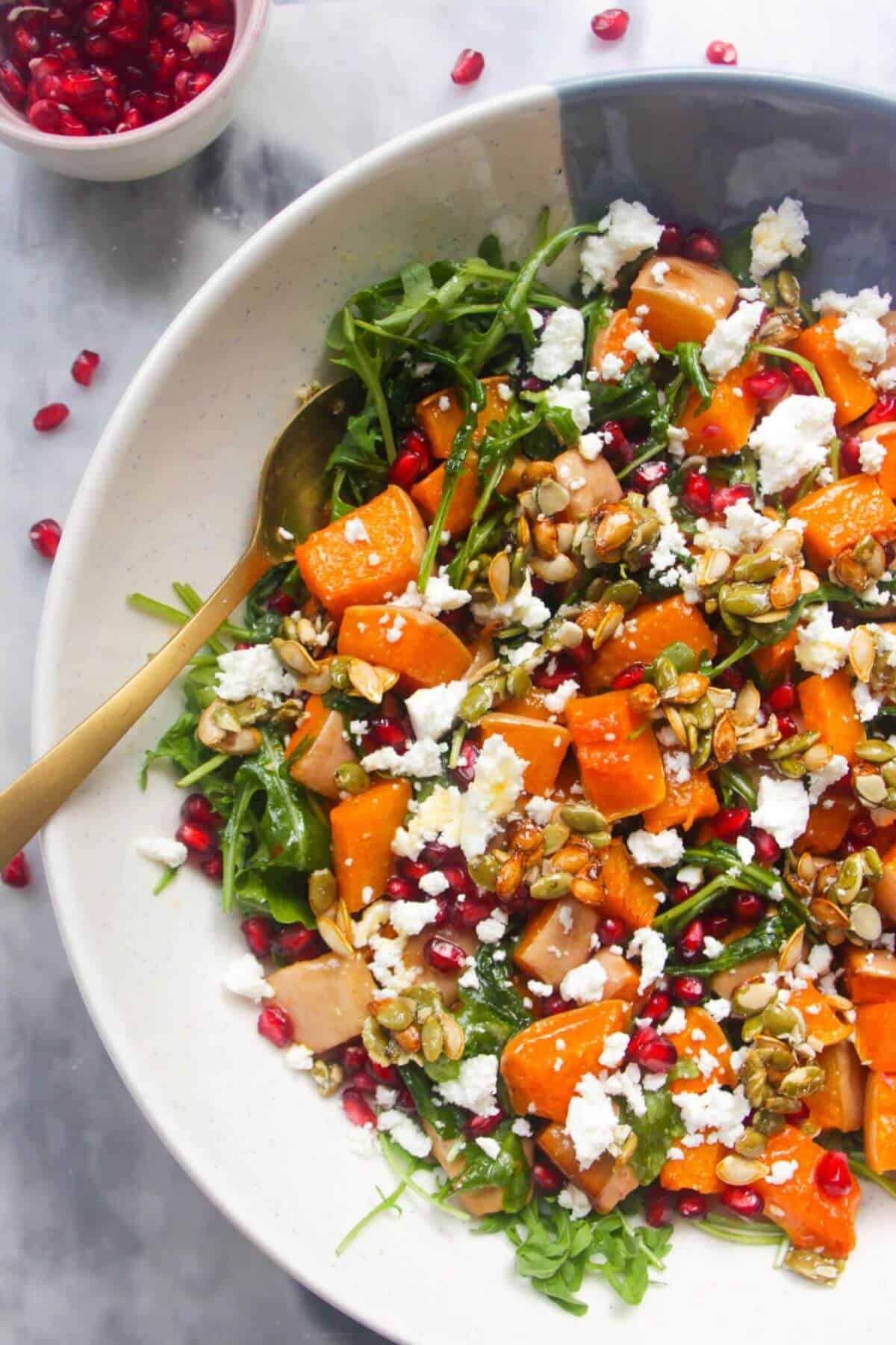 Large white and blue serving bowl with colourful butternut squash salad inside and pomegranate seeds on the side.