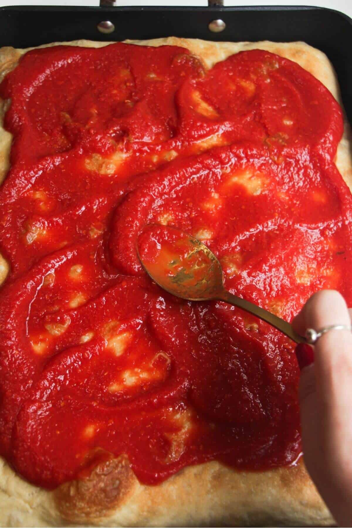 Spreading pizza sauce on top of par-baked focaccia in a large pan.