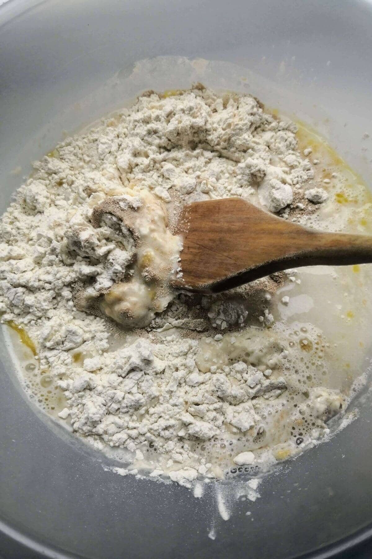 Foccacia dough about to be mixed in a large clear bowl with a wooden spoon