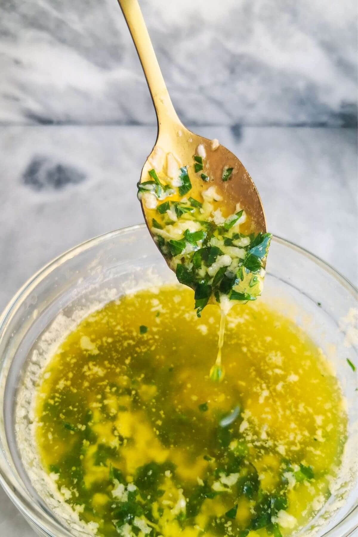 Garlic butter sauce in a small glass bowl, with a gold spoon with sauce on it, on a grey marble background.