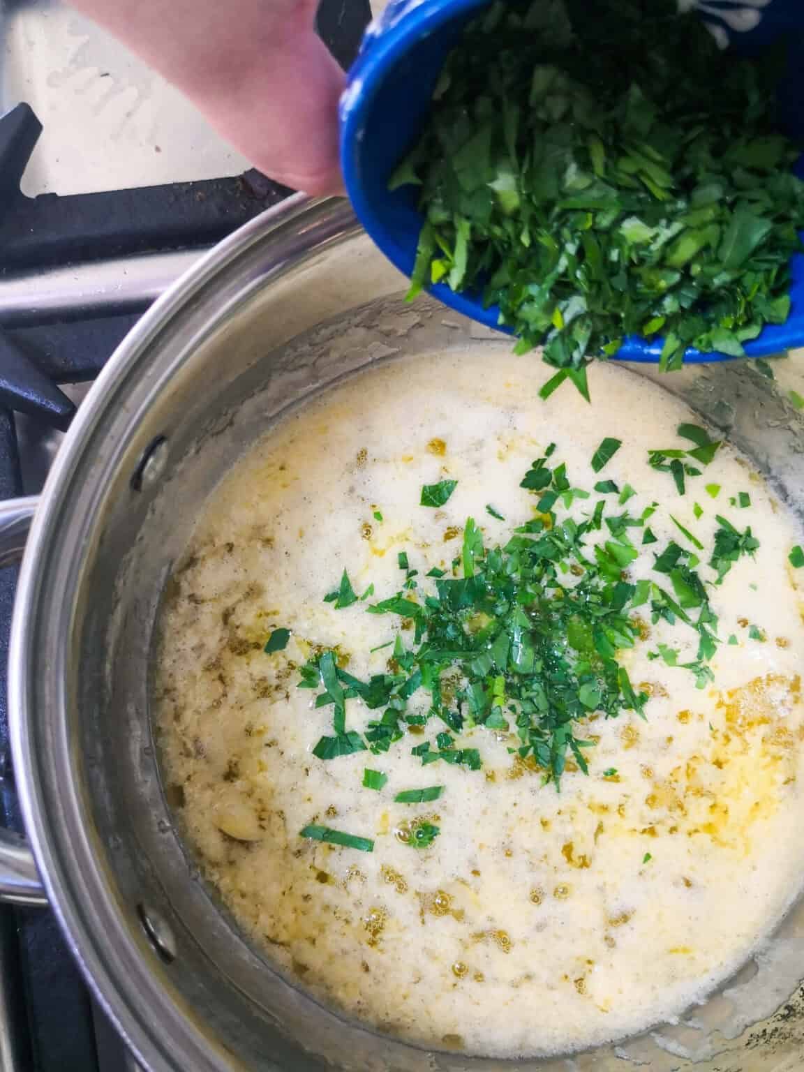 Chopped parsley being added to the garlic butter sauce in a small silver pan on the stove top.