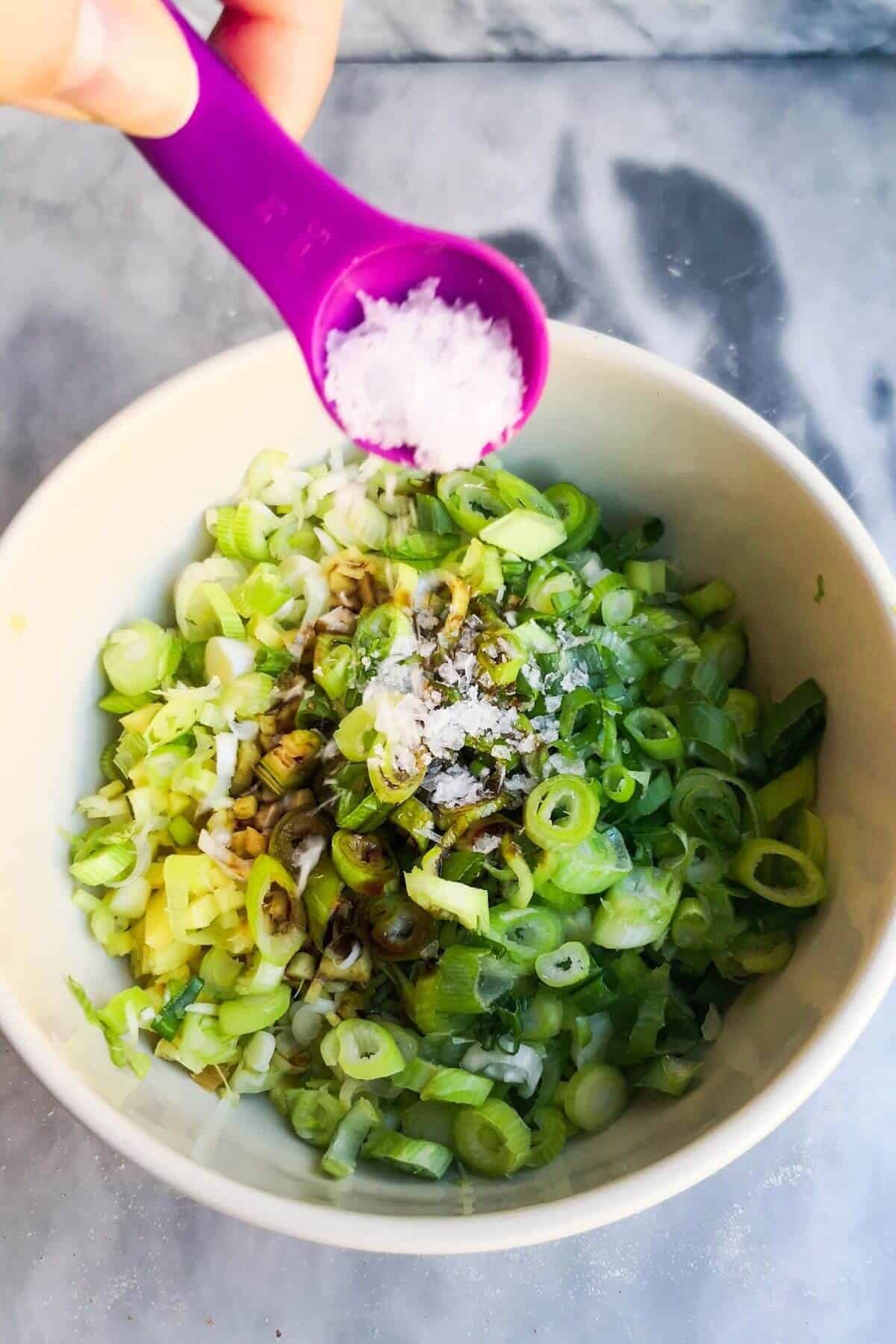 Small purple spoon pouring flaky salt into bowl with spring onion and ginger oil.