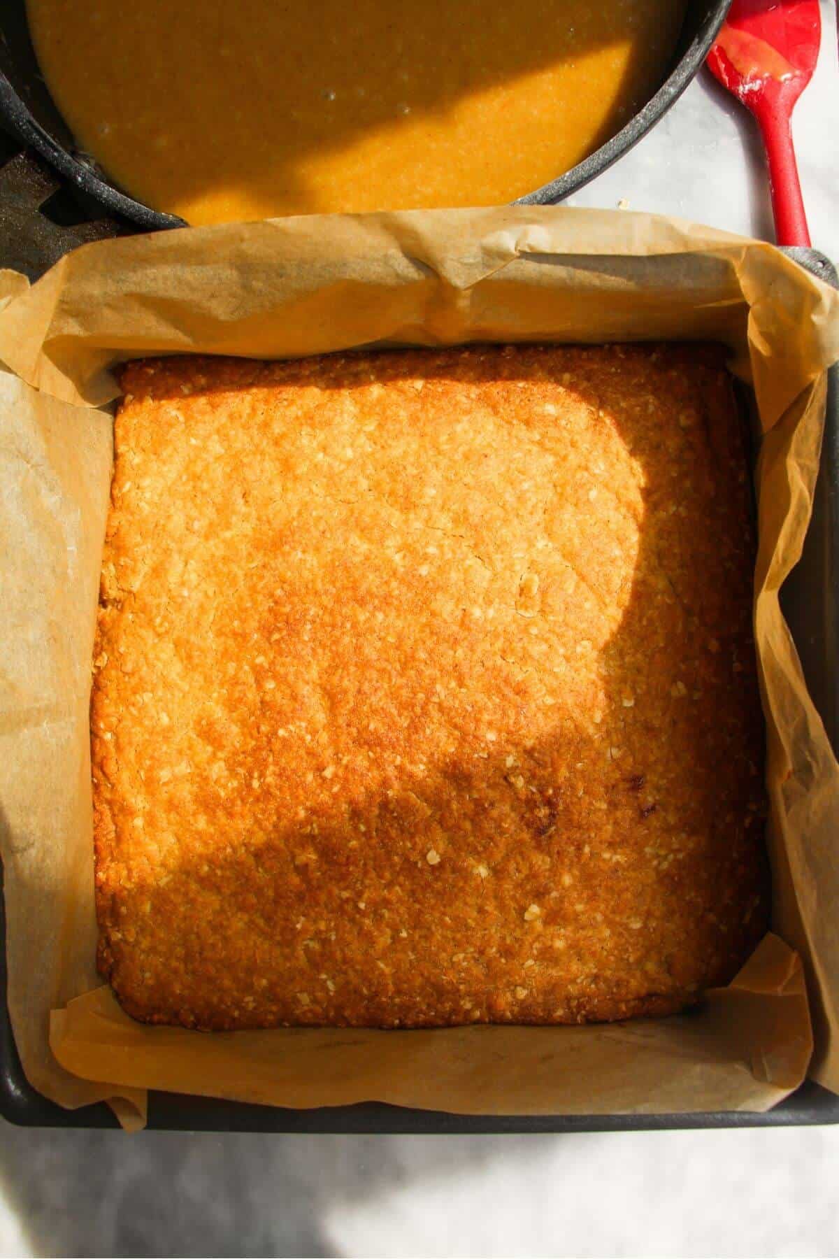Golden baked ginger crunch base in a lined baking tin with ginger icing in a pan in the background.