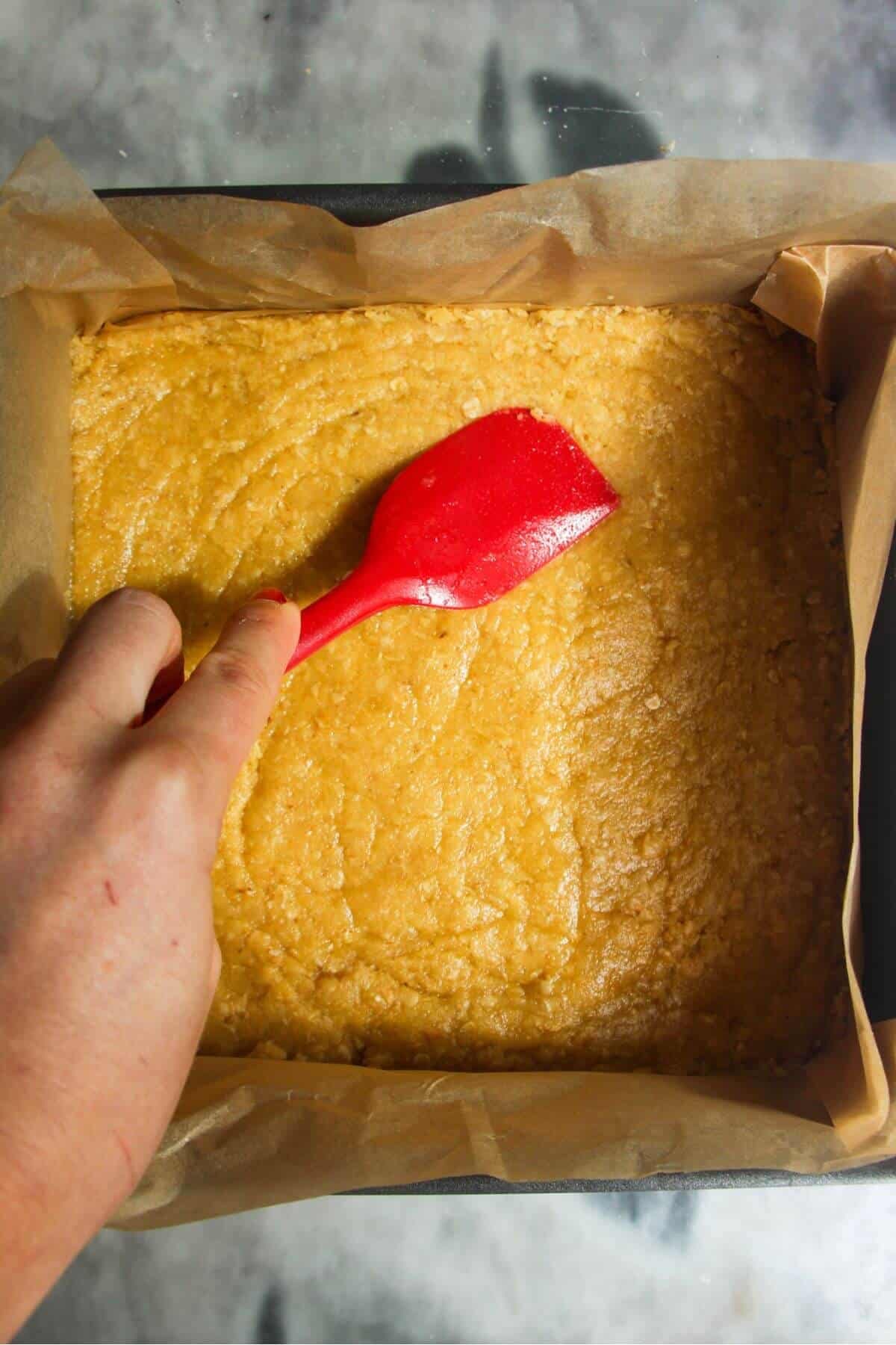 Red spatula smoothing biscuit base into lined baking tin.