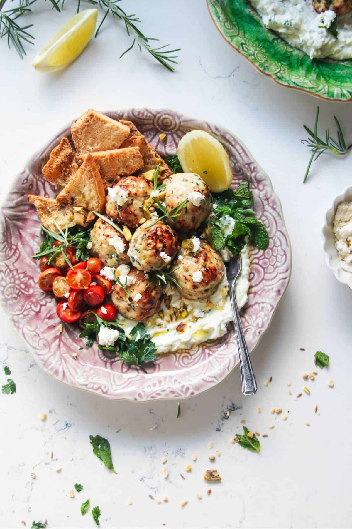 Chicken meatballs, tomatoes, on tzatziki with pita chips on a pink plate.