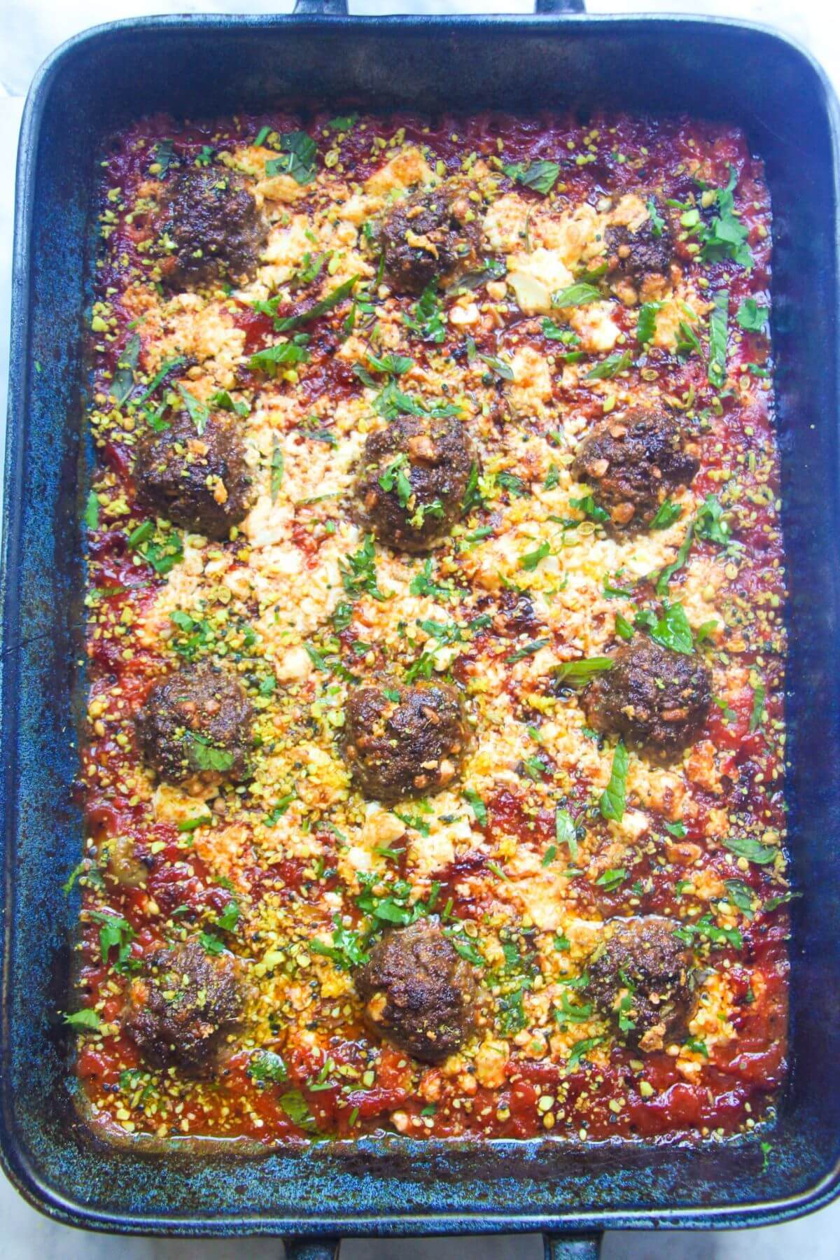 12 cooked meatballs in tomato sauce, topped with feta and green herbs in a large blue oven dish.
