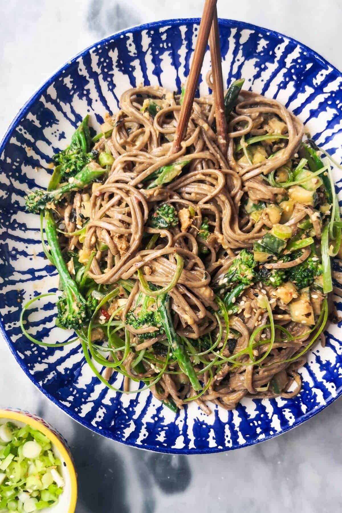 Sesame soba noodles with cucumber and broccolini in a white and blue bowl with chopsticks, with a small bowl of spring onions on the side.