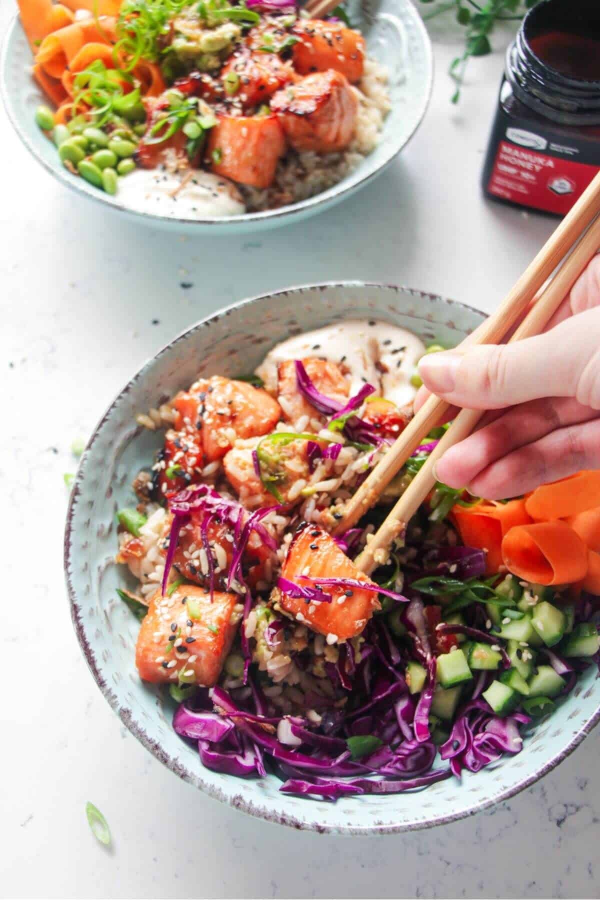 Hands holding chopsticks, picking up piece of salmon from poke bowl.
