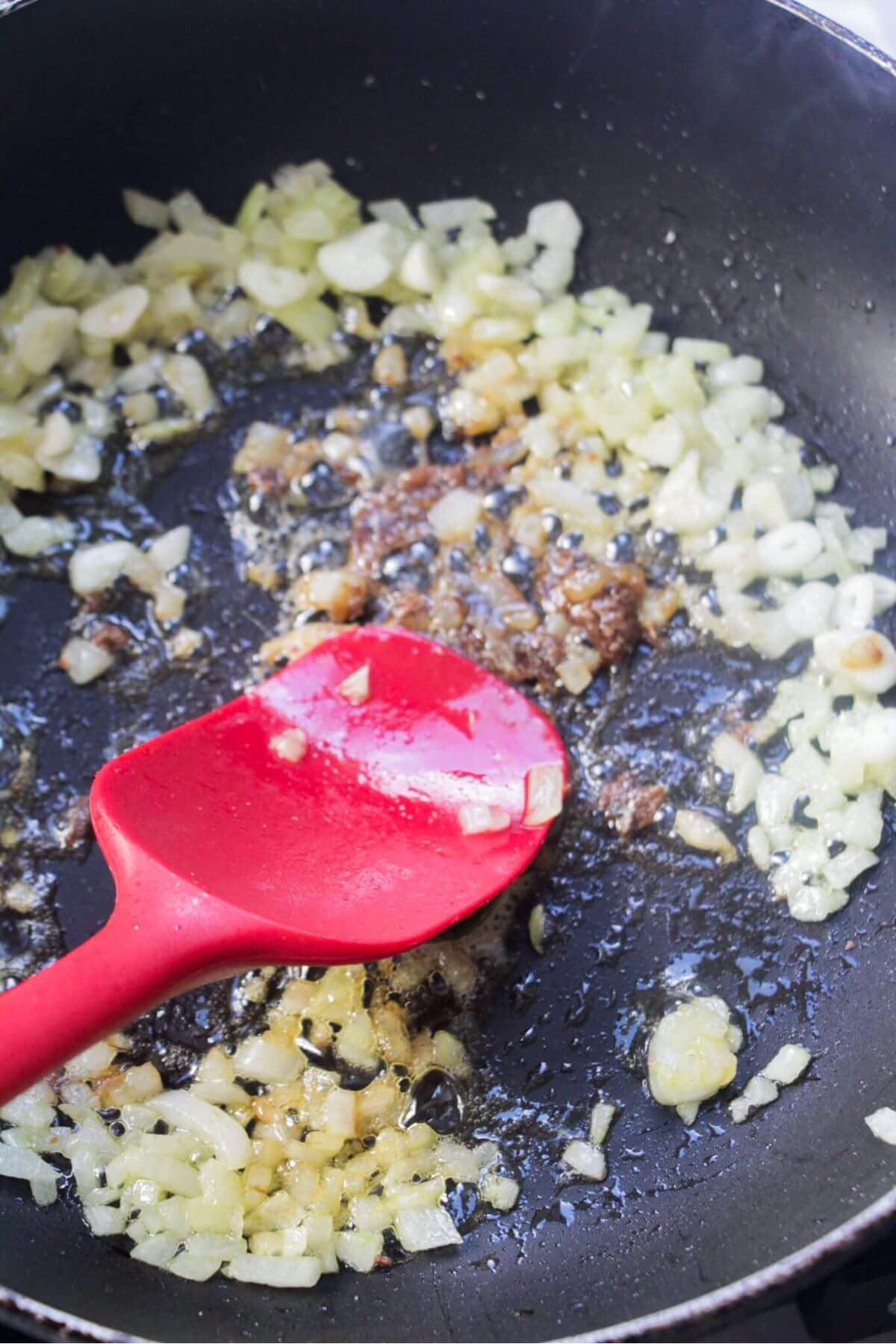 Softened onions, garlic and anchovies in a small black pan with a red spatula squashing the anchovies into the oil.