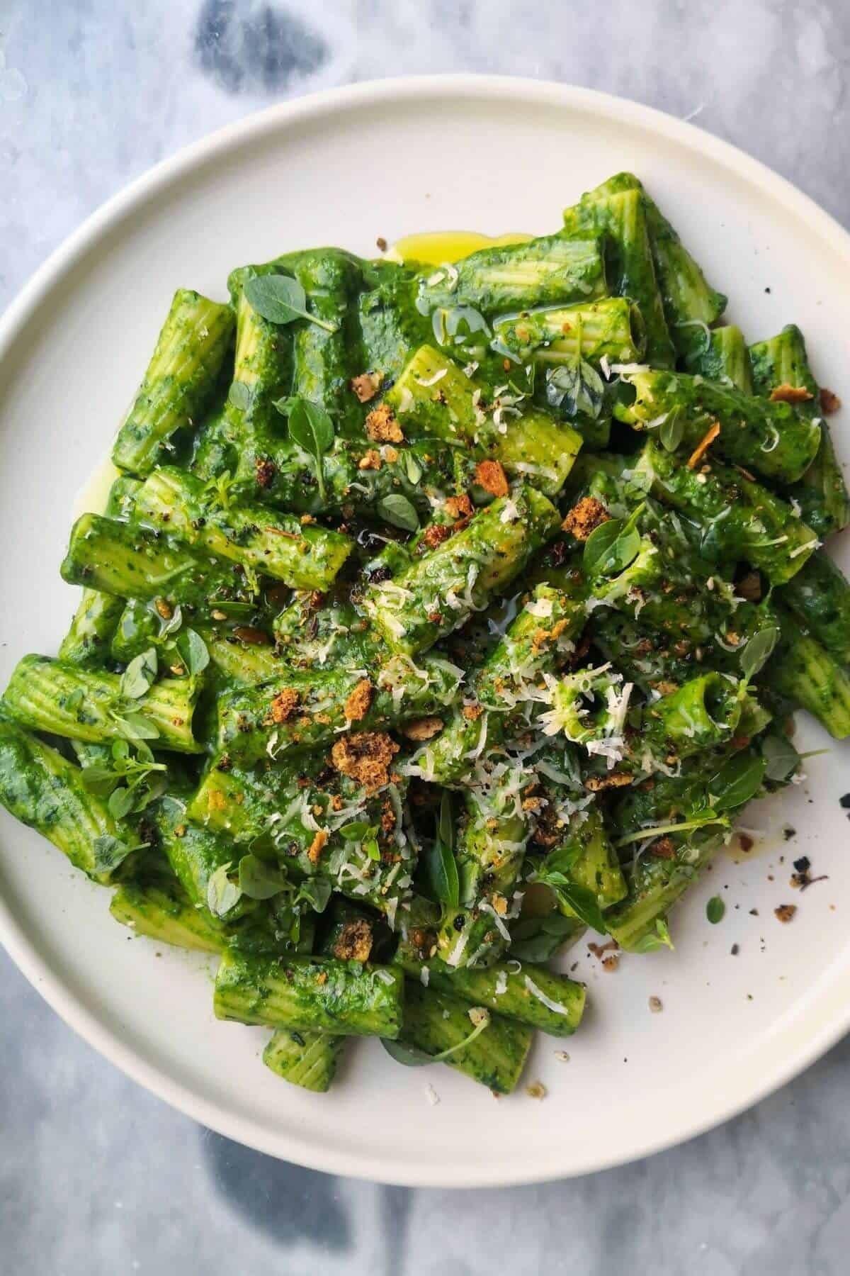 Green pasta sauce with rigatoni on a white plate, topped with grated parmesan, chilli oil and crispy pita crumbs.