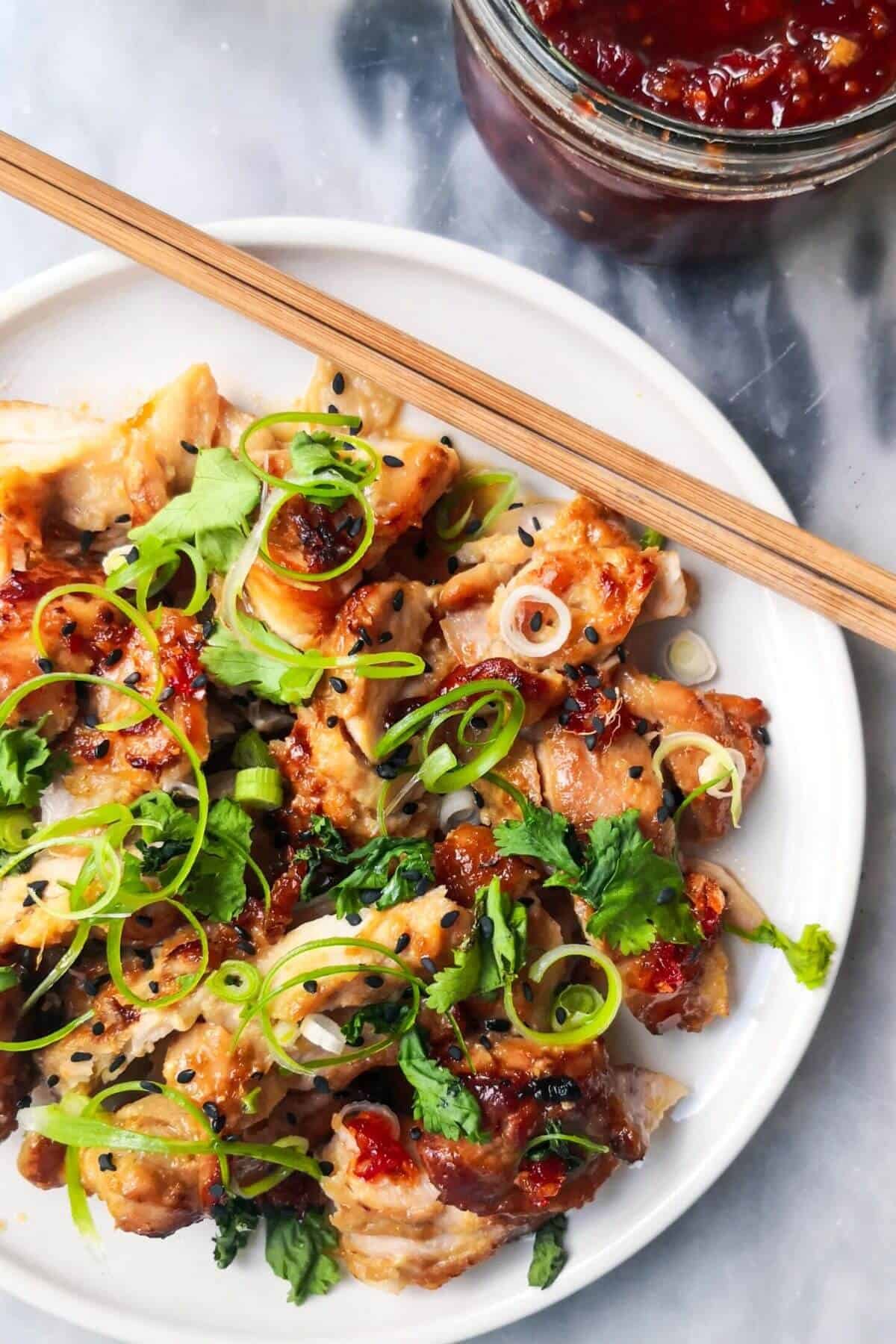 Miso chicken on a white serving plate with spring onion, coriander, black sesame seeds with chopsticks on the plate and a jar of chilli jam on the side.