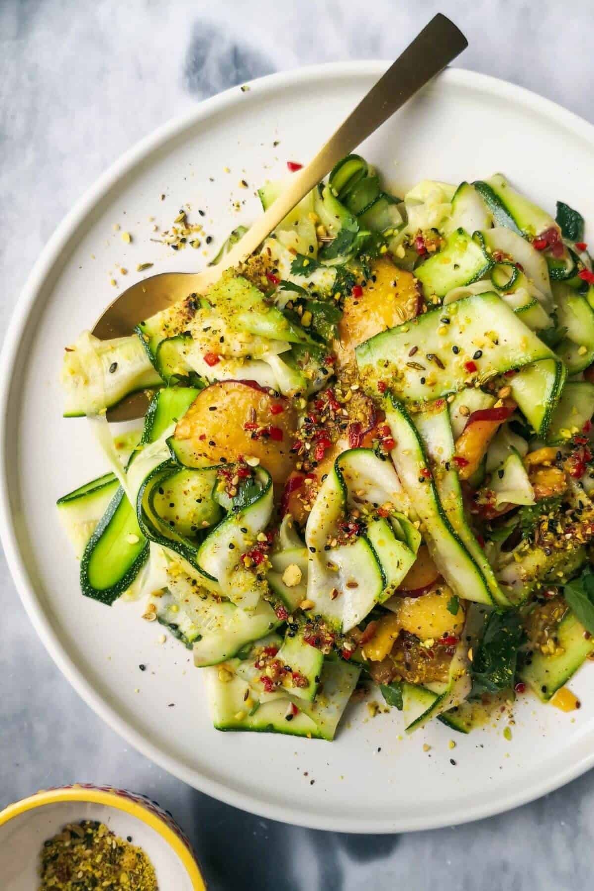 Raw courgette and peach salad on a white serving plate on a grey marble background with a small bowl of dukkah on the side.
