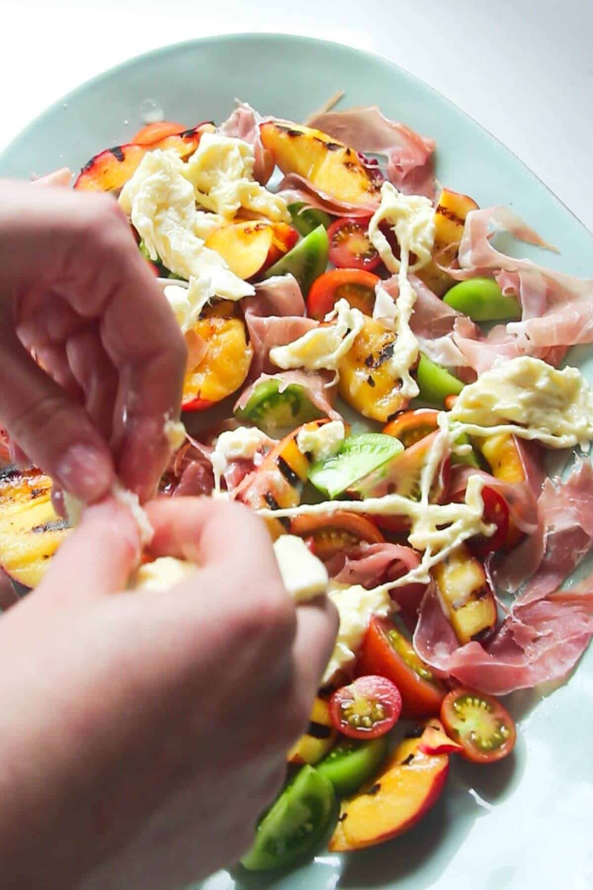 Hand tearing burrata on top of peaches, tomatoes and prosciutto on a large blue oval plate.