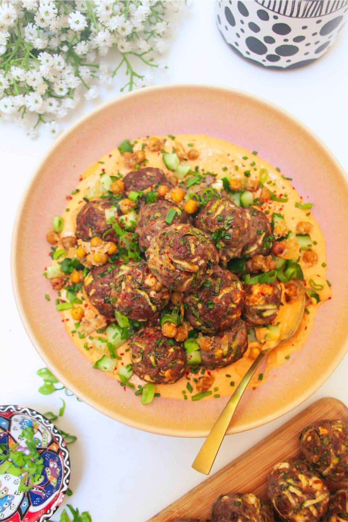 Meatballs piled on top of smoky red pepper sauce with flowers in the background.