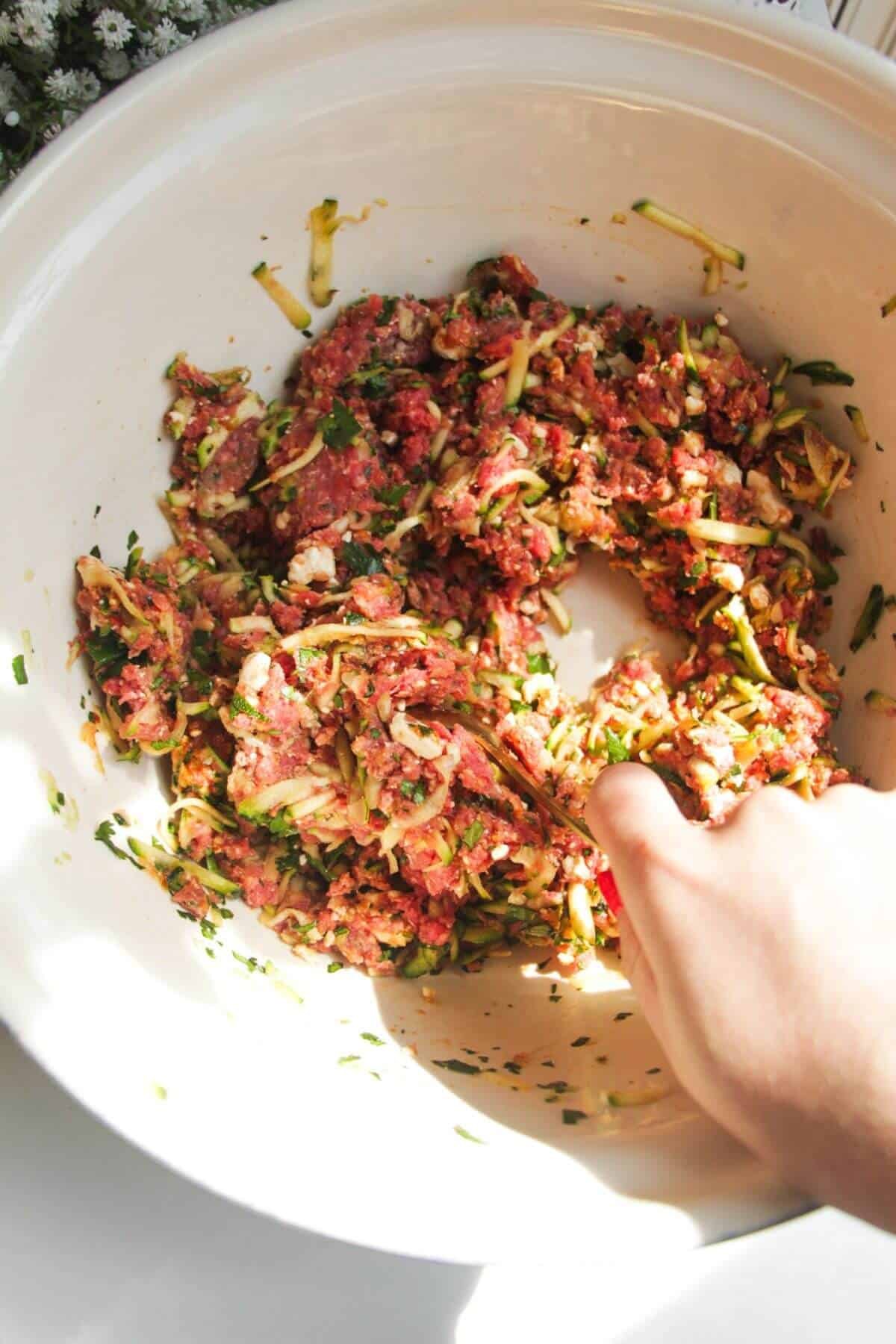 Hand holding a spoon mixing zucchini, feta meatball mixture.