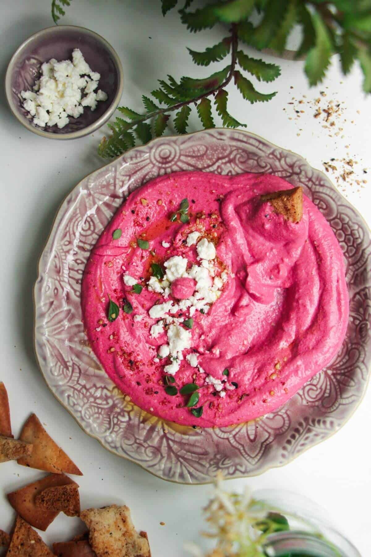 Bright pink beet feta dip on a pink plate with feta crumbled on top and pita chips on the side.