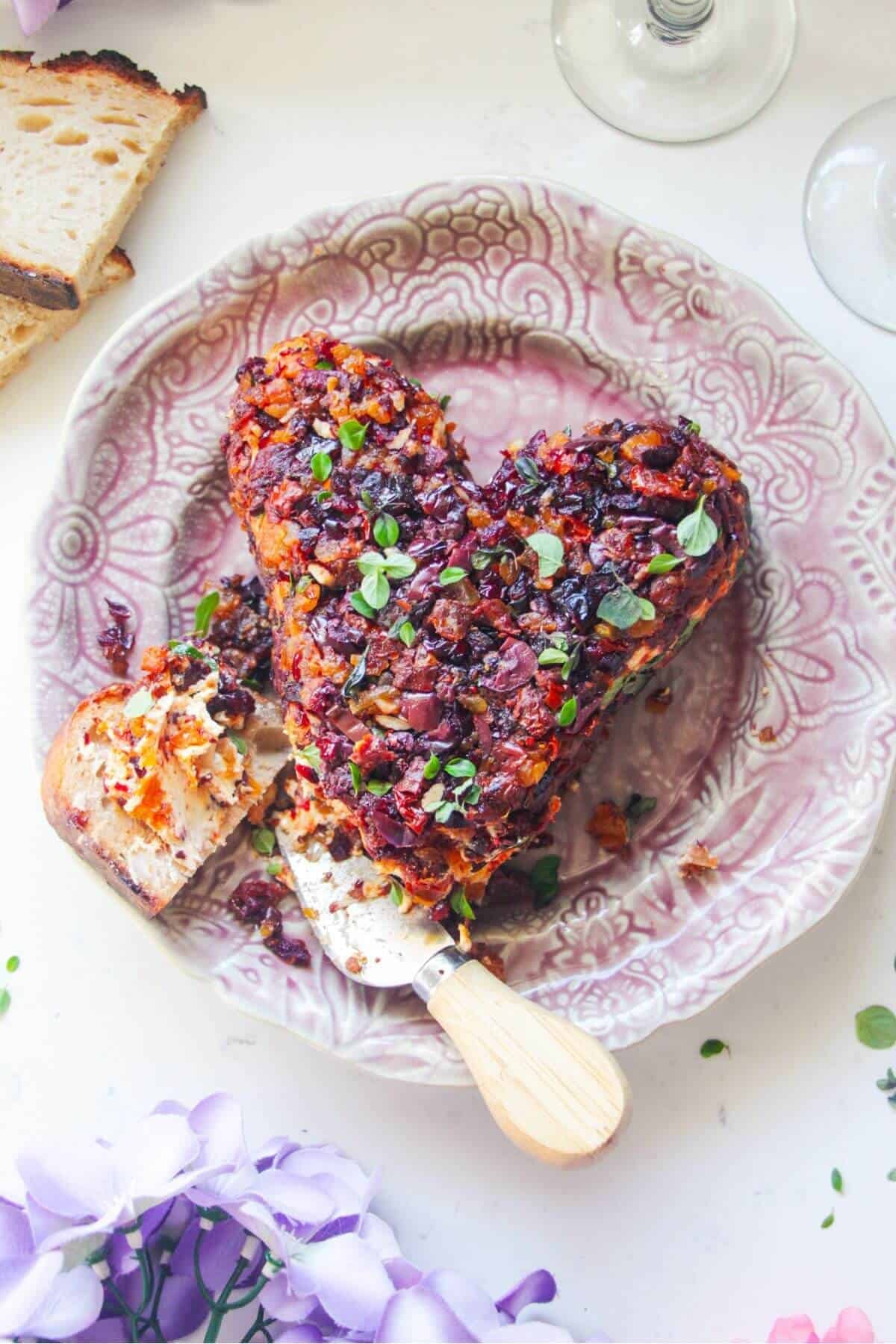 Heart shaped cheese ball, covered in dried fruit on a small pink plate, with bread on the side.