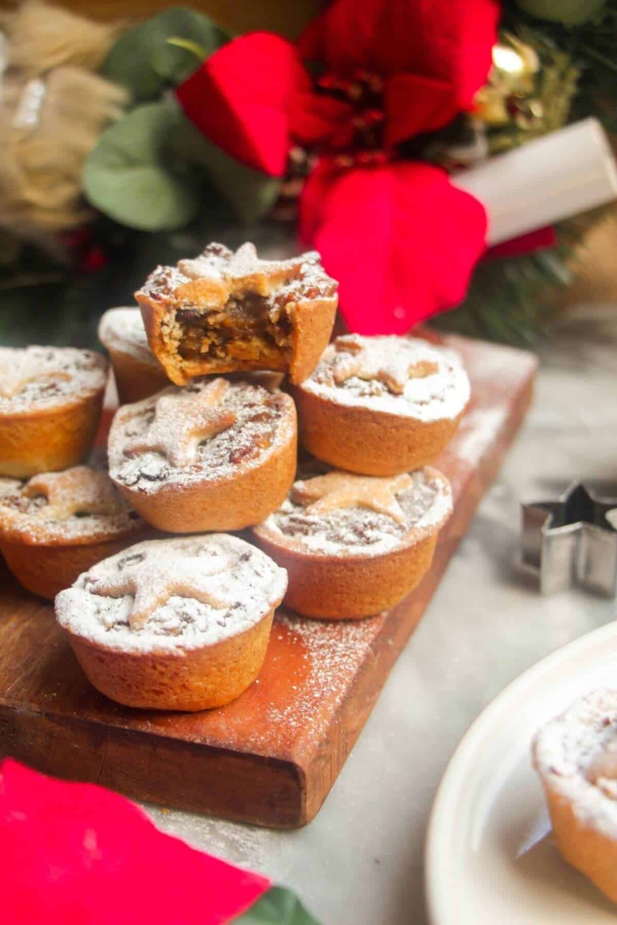 A pile of icing sugar dusted Christmas mince pies on a small wooden board with Christmas deocrations in the background.
