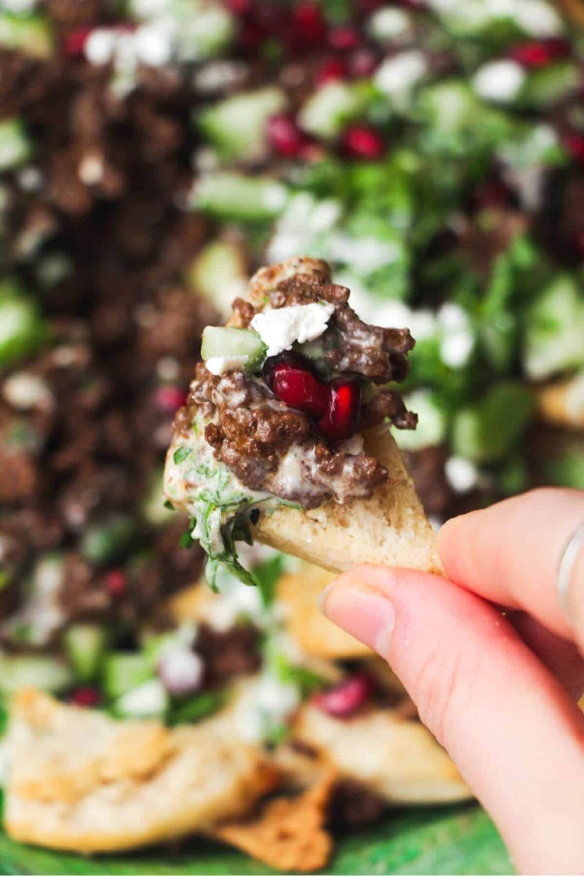 Close up of a hand holding up a loaded pita chip with beef and pomegranate seeds.
