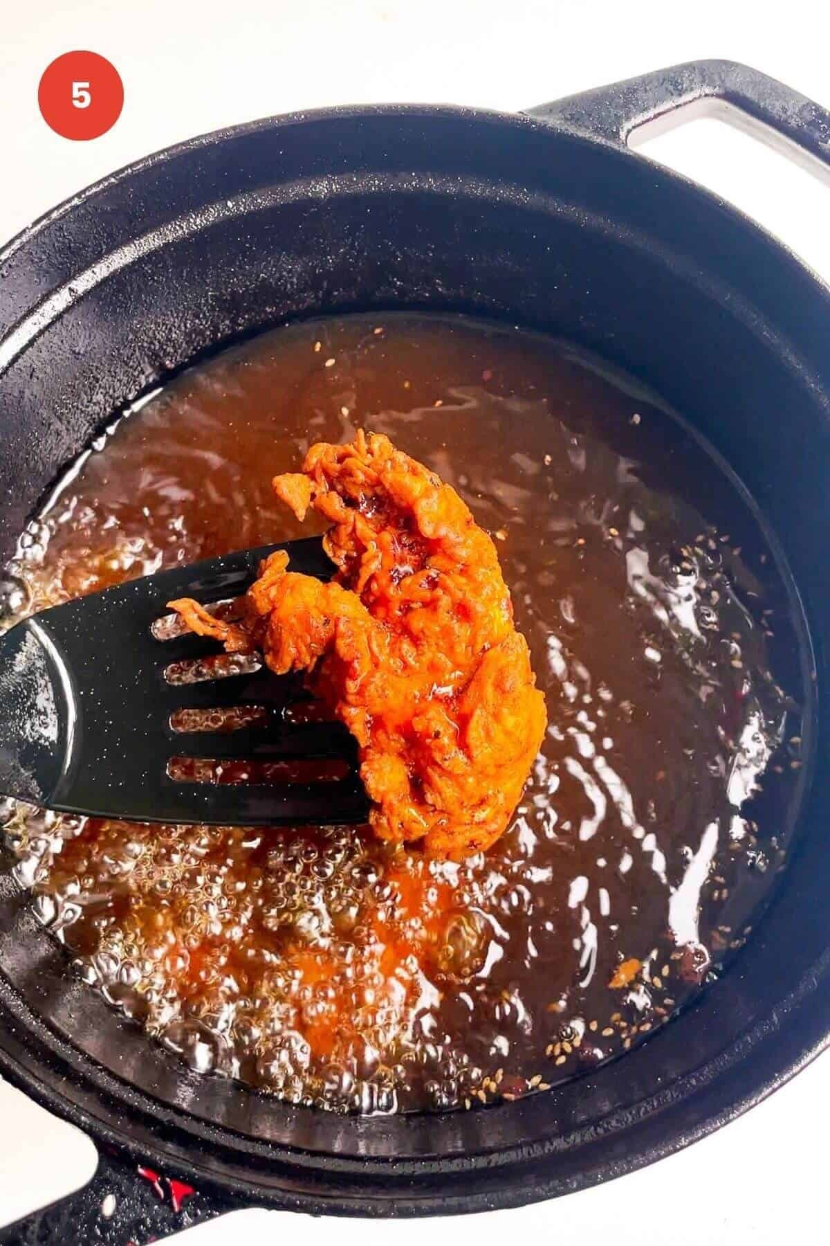 Spatula holding up fried chicken tender with pot of chicken frying behind.