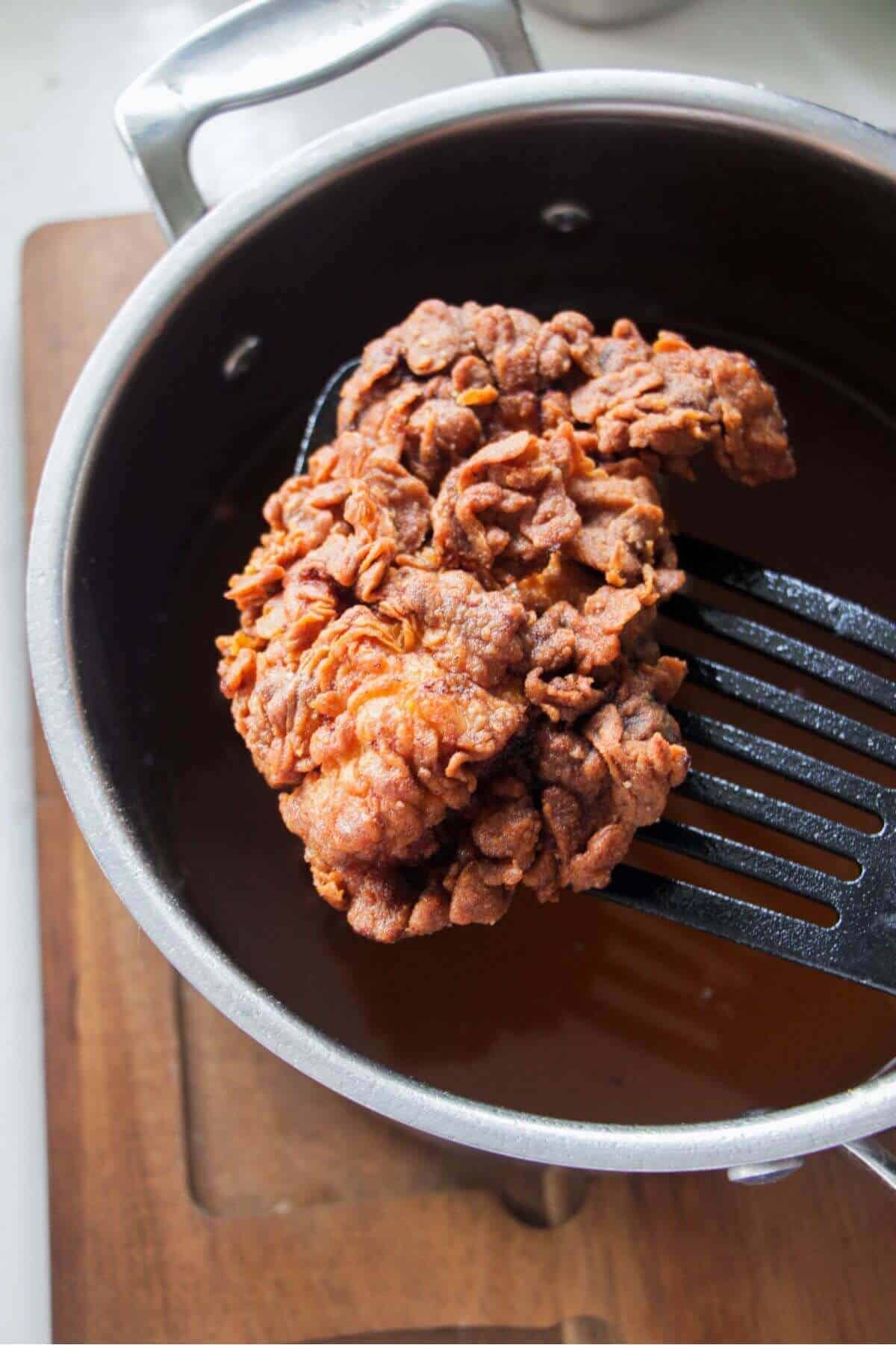 Piece of fried chicken on a black spatula with pot of oil in the background.