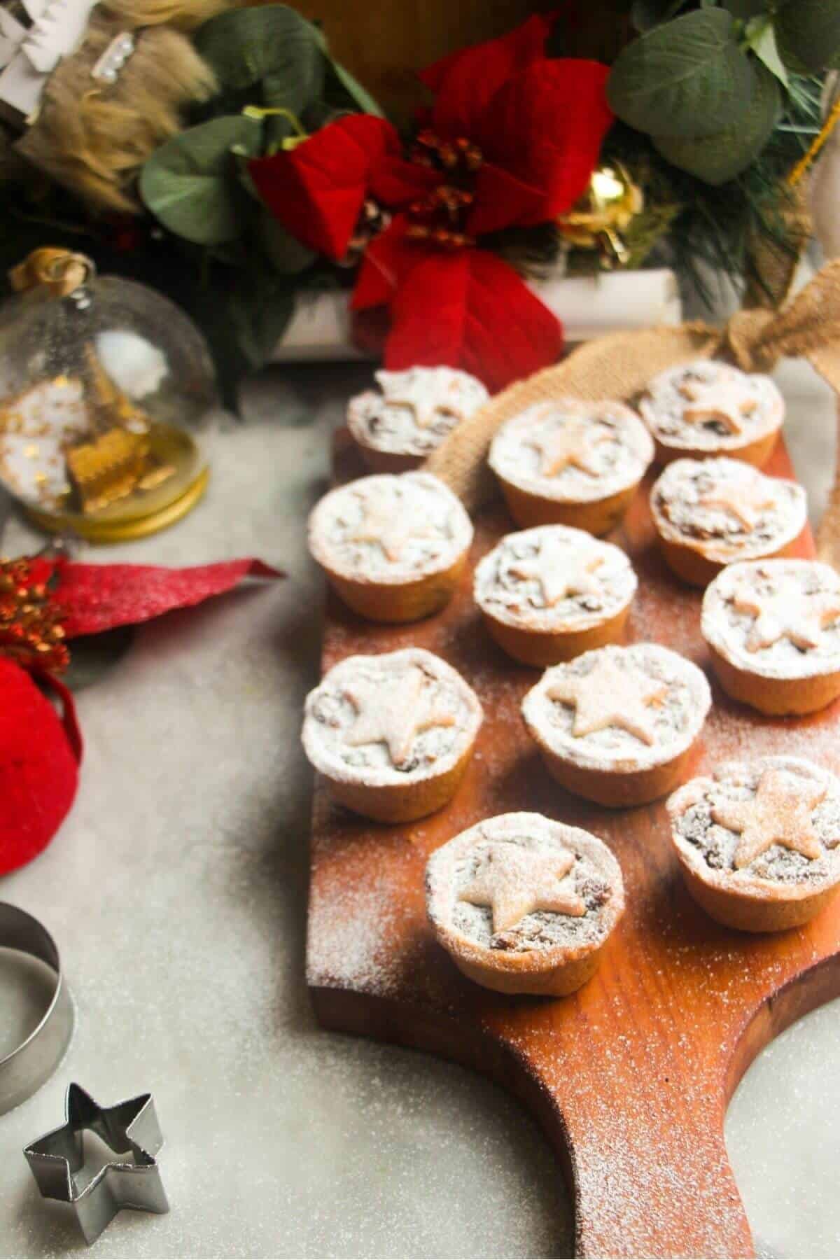 Icing sugar dusted Christmas fruit mince pies on a small wooden board with red and gold decorations in the background.