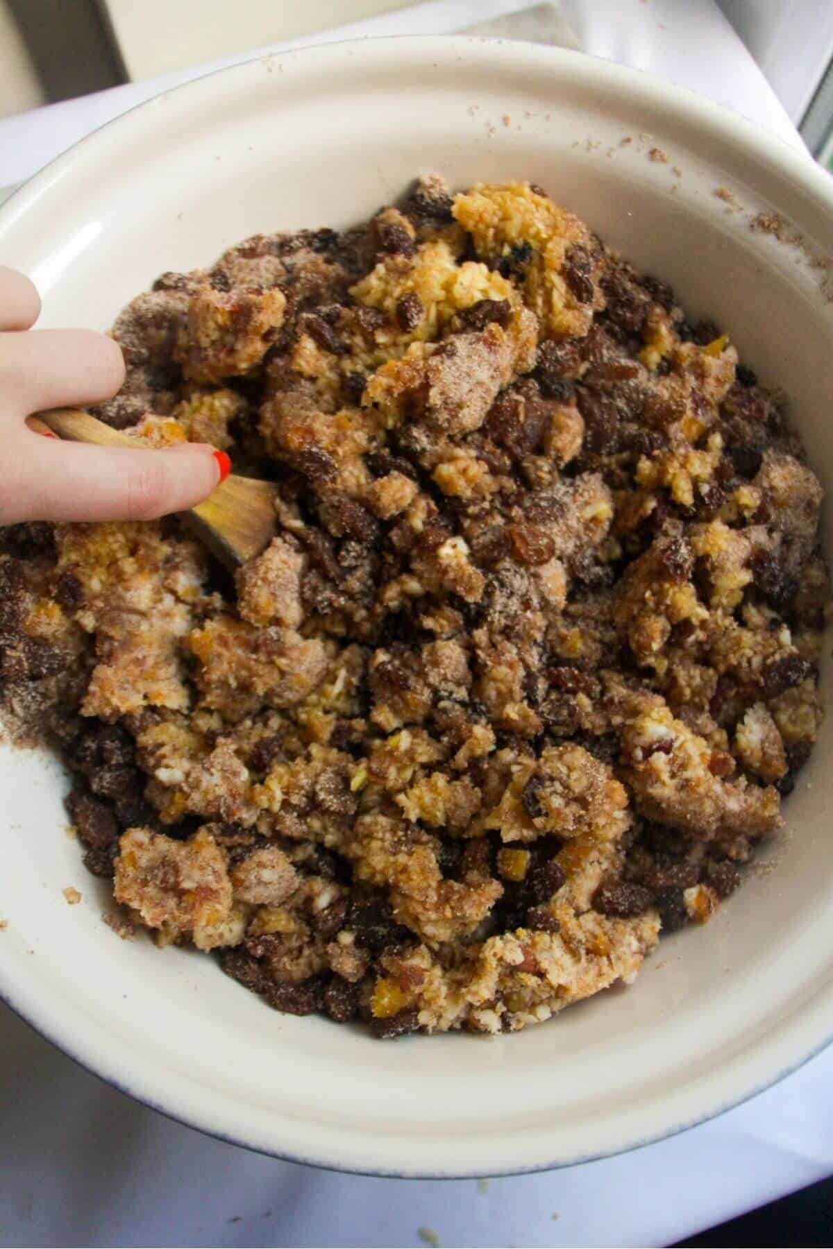 Hand holding a wooden spoon and stirring Christmas fruit mince in a large white bowl.