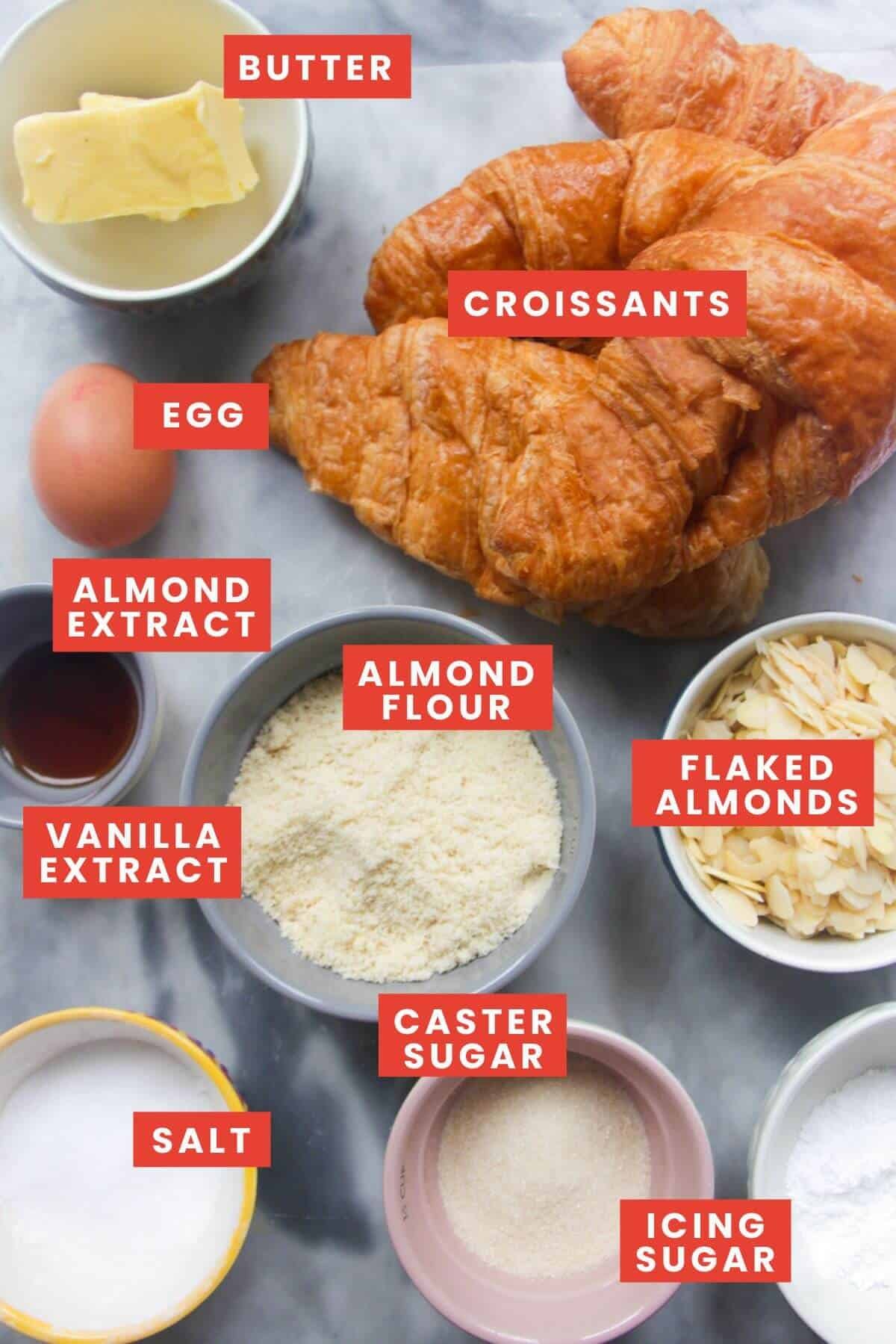 Ingredients for almond croissants laid on a grey marble background and labelled.