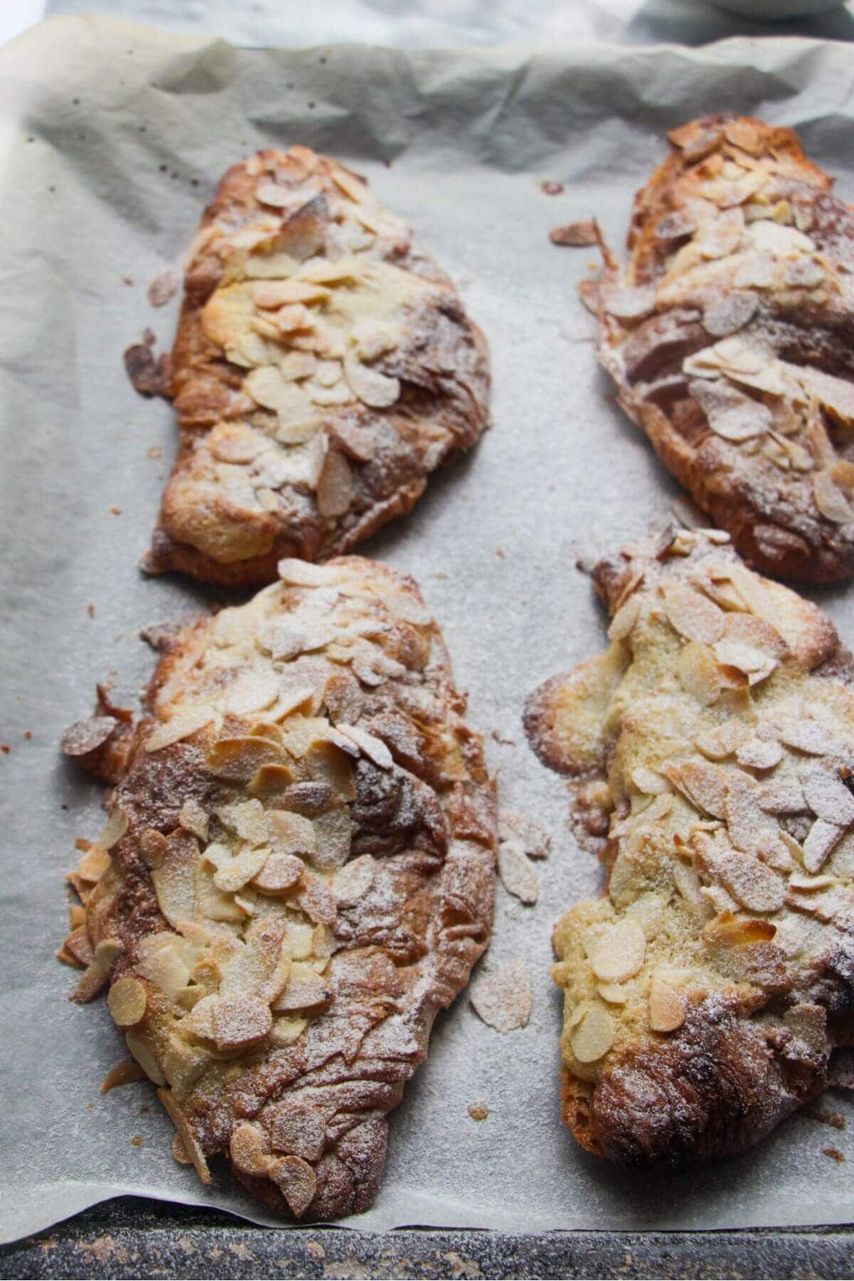 Four cooked almond croissants on a lined baking tray, with icing sugar on top.