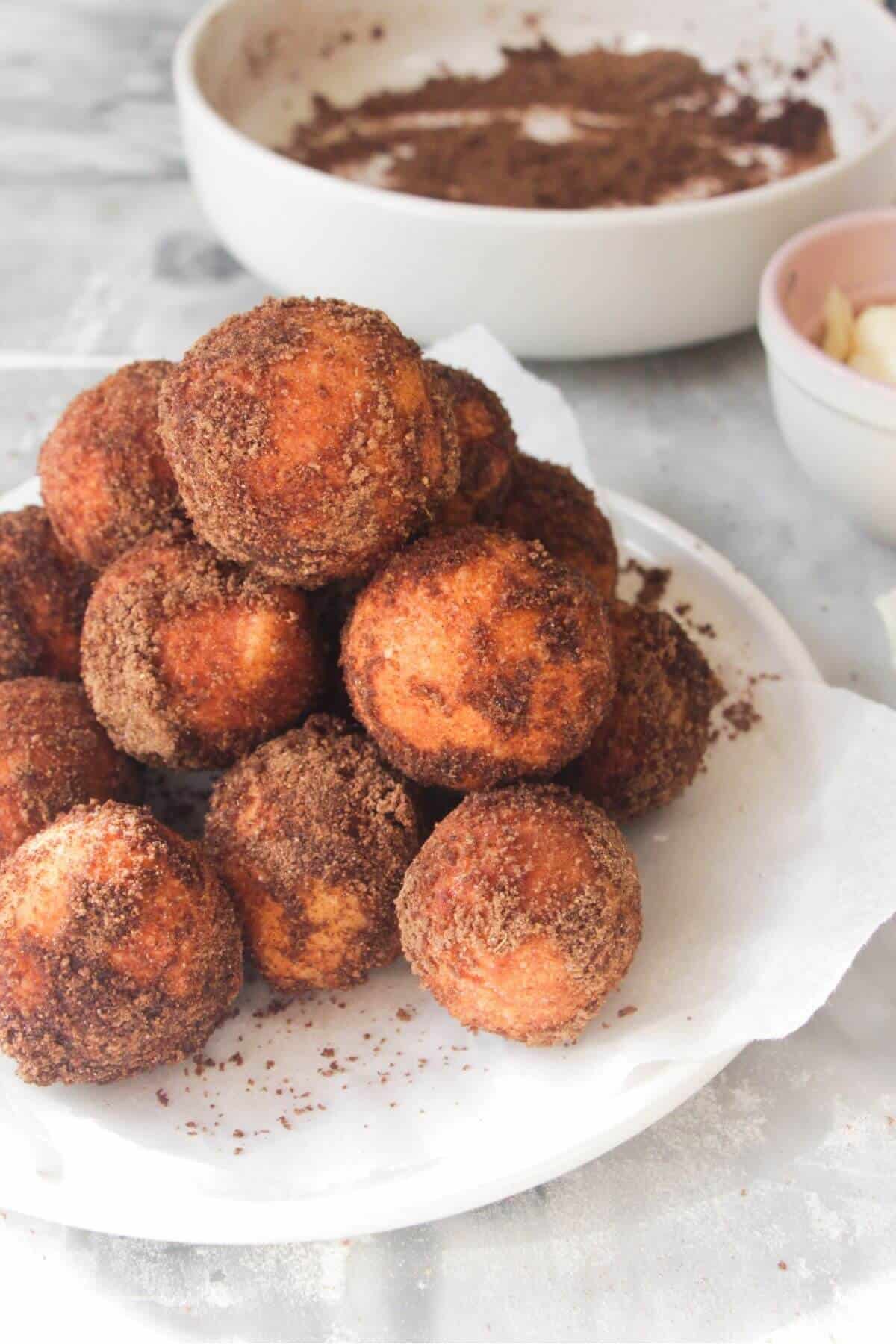 A pile of sugar coated donut bites on a small white plate, with bowl of more pumpkin spice sugar in the background.