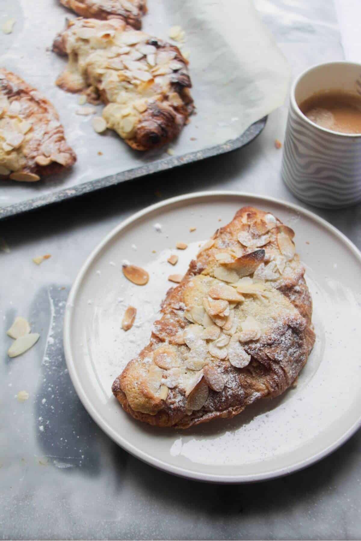 An almond croissant on a small white plate with a tray of almond croissants in the background.