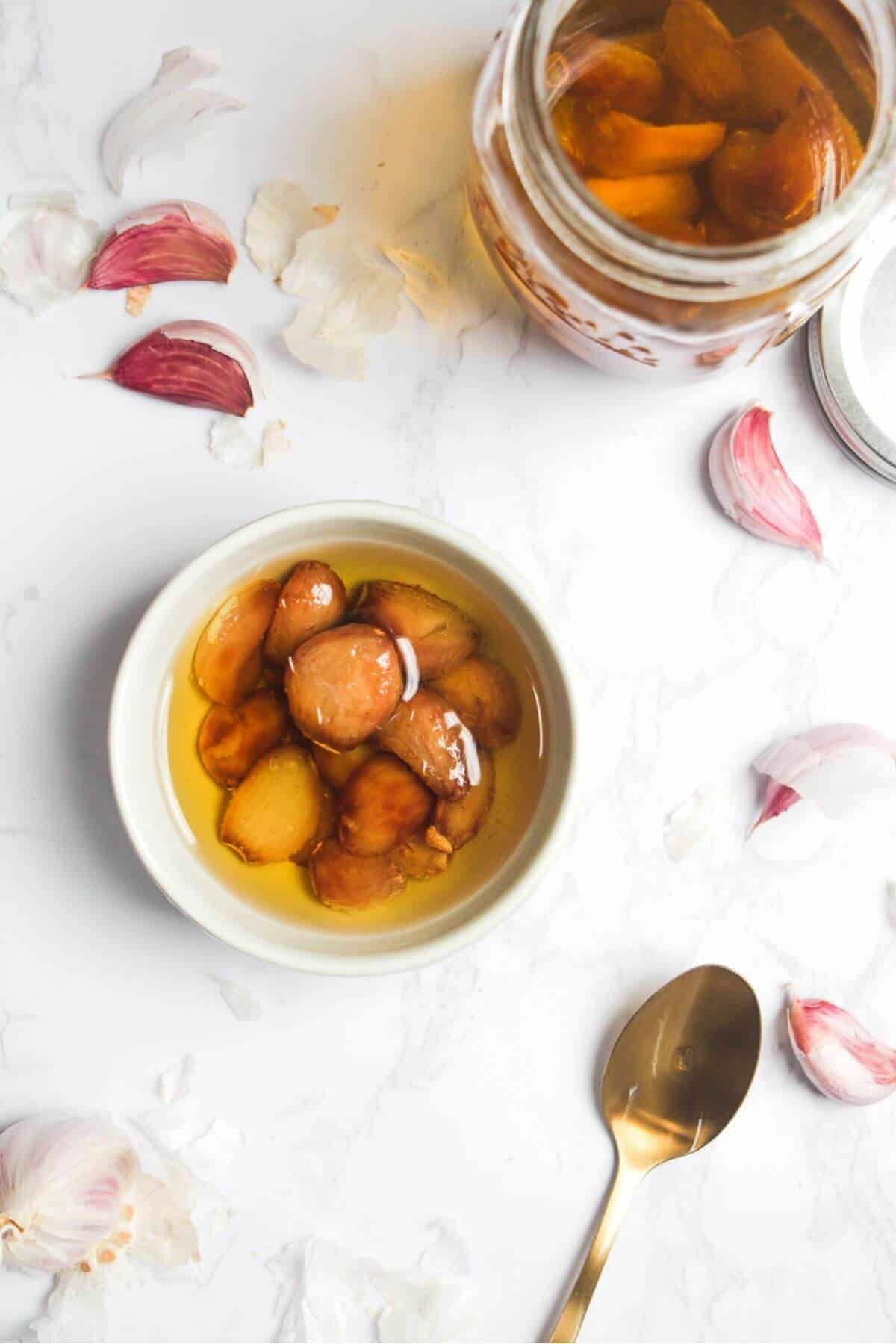 Garlic confit in a small white bowl with a gold spoon on the side, and a jar of garlic confit in the background.