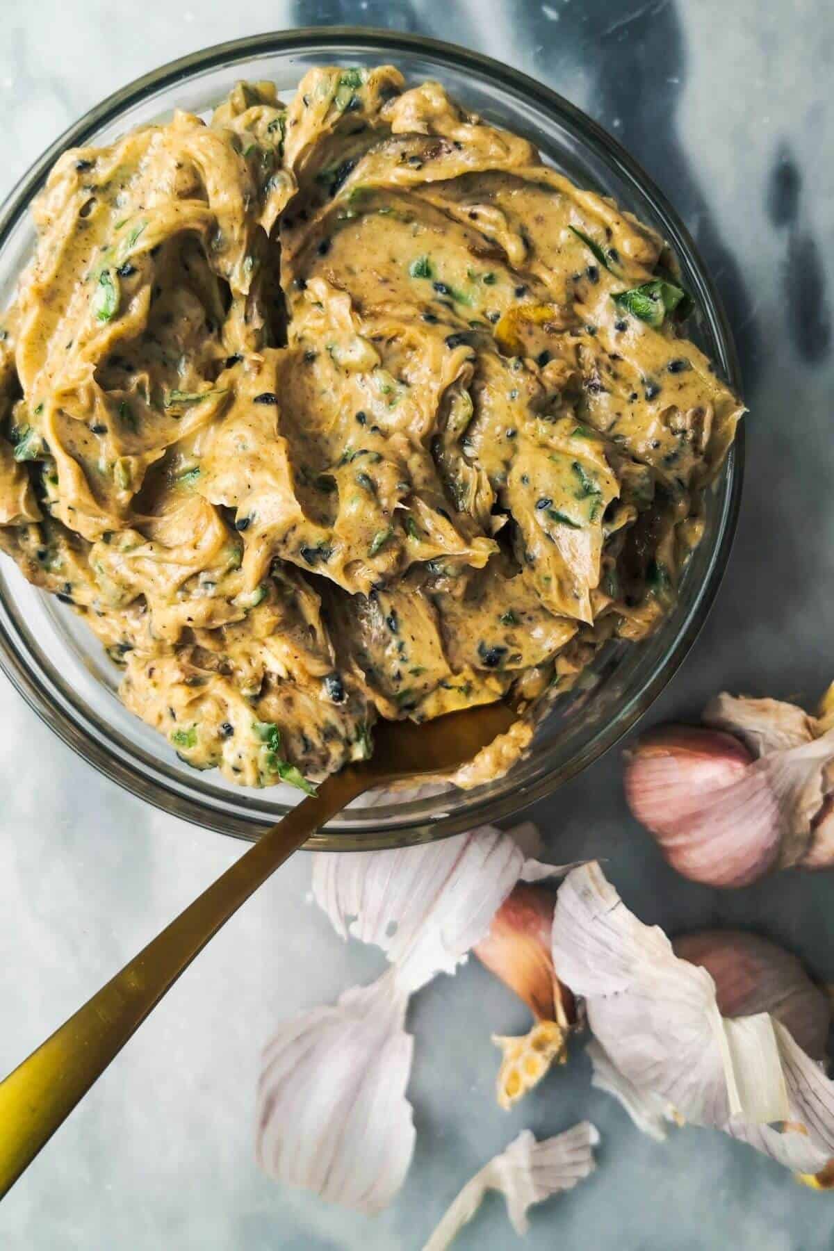 Roasted garlic butter in a small glass bowl with a gold spoon inside, with garlic cloves in the background.